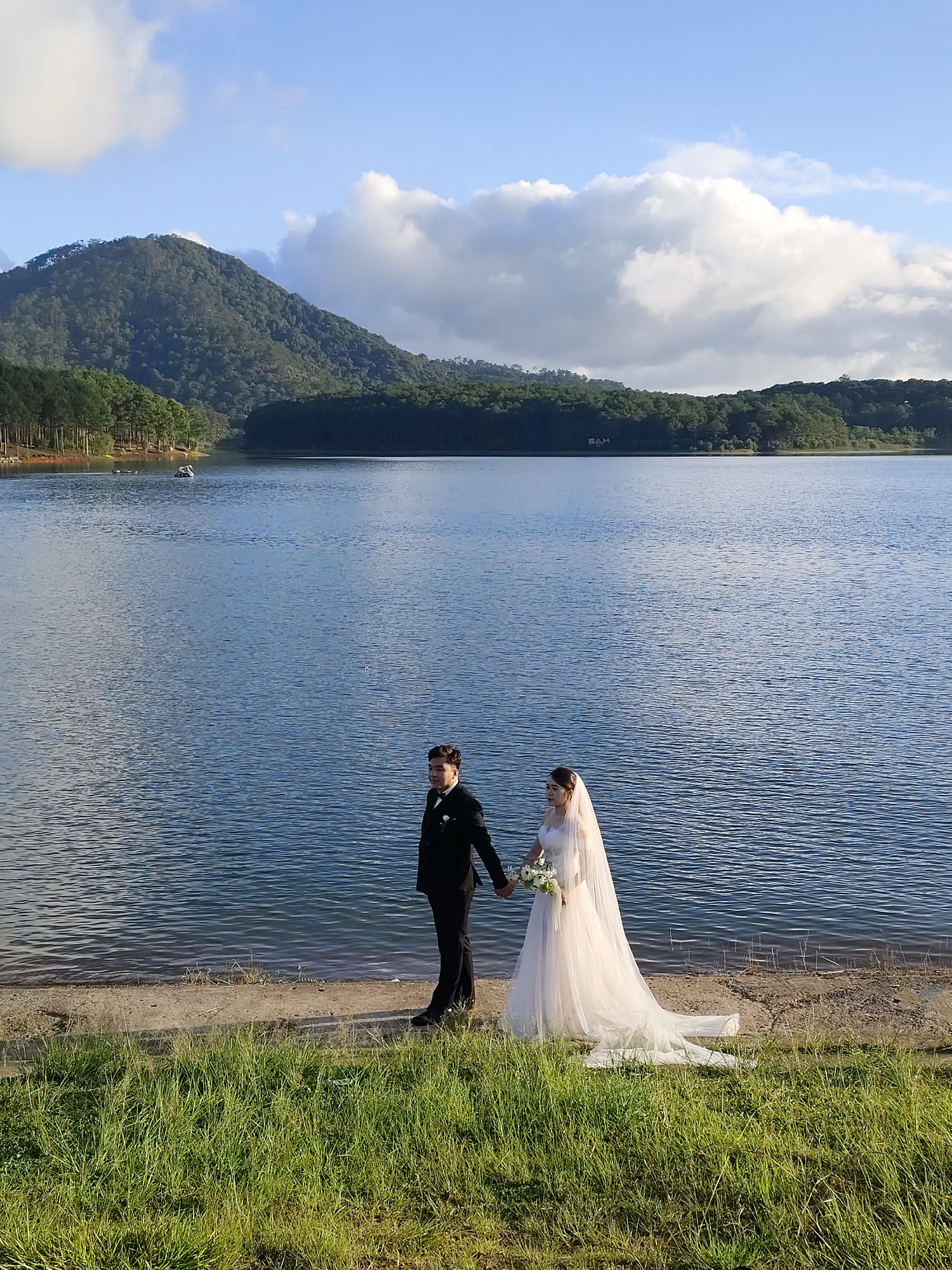 A wedding photo captures a bride and groom standing on the shore of Tuyen Lam Lake, located in Da Lat, Vietnam. The bride is wearing a white wedding dress with a long train and veil. She holds a bouquet of white flowers. The groom is in a black suit. They are holding hands.
Behind them is the lake and a mountain covered with trees. A few clouds are scattered across the blue sky. The shoreline is composed of grass, sand, and a small concrete portion.