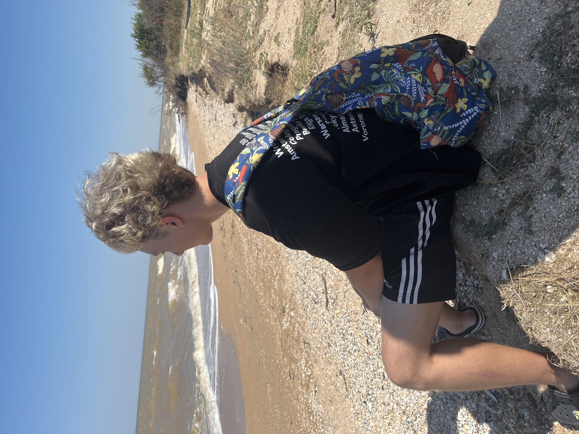 A young male with blond, curly hair sits on a sandy beach in Dolzhanskaya, Russia, looking out at the Sea of Azov. The sky is clear and blue. The waves are choppy, indicating a breezy day. The beach is covered in sand and shells. The male is wearing a black t-shirt with text printed on the back and black athletic shorts with white stripes. A backpack with a colorful floral pattern is slung over his right shoulder. His left leg is visible, showing a light scar on the shin. He is wearing black Crocs-style sandals. In the background, dry vegetation and sand dunes line the coast. The image captures a moment of quiet contemplation by the sea.