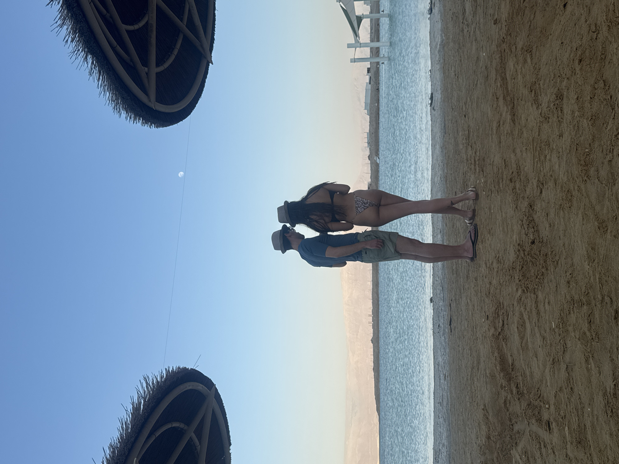 A man and a woman stand facing away from the camera on a sandy beach, looking out at a body of water. The man is wearing a blue t-shirt, khaki shorts, and a hat. The woman is wearing a bikini with a tiger-striped pattern and a hat. The water is calm and reflects the clear blue sky. In the distance, there are low, barren hills. Two thatched umbrellas provide shade on the beach. The overall scene suggests a relaxed vacation setting.