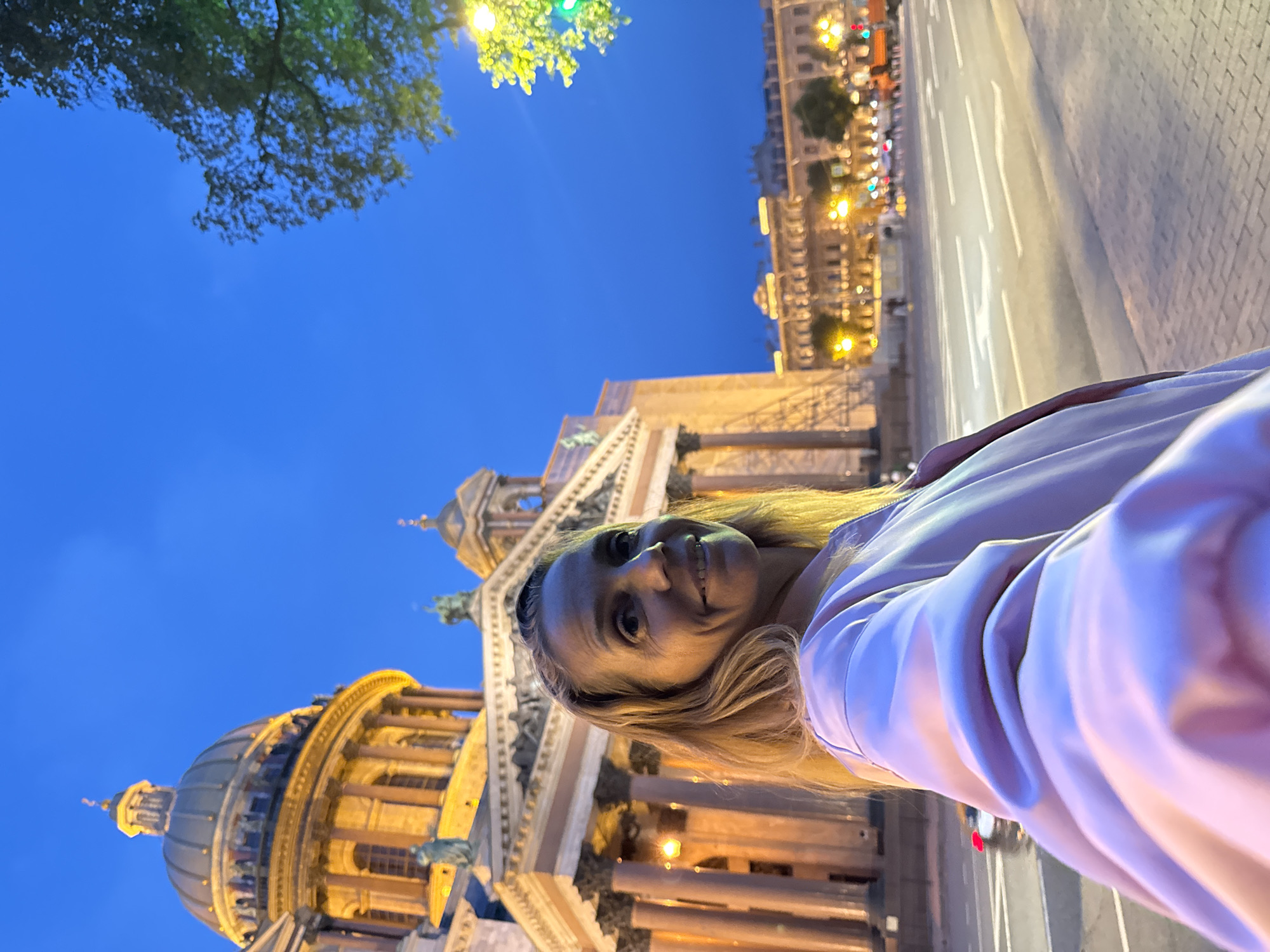 A woman is taking a selfie in front of a large, ornate building with a dome. The building is illuminated by warm, artificial lights, suggesting it is evening or night. The sky is a deep blue, indicating dusk or nighttime. A street with traffic lights and other illuminated buildings is visible in the background. A tree with green leaves is in the upper left corner, adding a touch of nature to the urban scene. The woman has blonde hair and is smiling, appearing happy and relaxed. She is wearing a light-colored jacket. The overall scene suggests a tourist experiencing a landmark in Saint Petersburg, Russia at night.