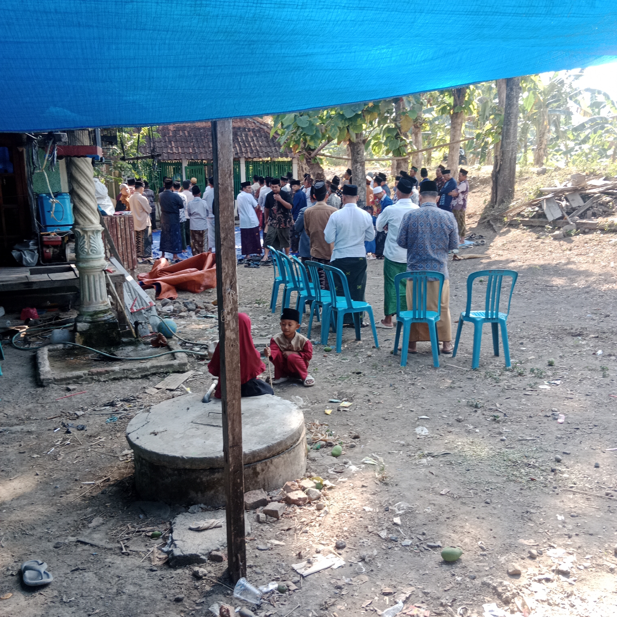 In Tegowanu Kulon, Indonesia, an outdoor gathering is taking place under a large blue tarp. A group of men, many wearing white shirts and dark peci hats, stand in clusters, some facing away from the camera. Several blue plastic chairs are arranged in rows. In the foreground, a young child wearing a red outfit and a black peci is sitting on the ground, holding a stick. A woman in a red hijab is seated behind the child. The ground is dirt and gravel, with scattered debris including a discarded flip-flop, plastic bottles, and natural detritus. A concrete structure, possibly a well or a planter, is visible in the lower left. In the background, trees and a building with a tiled roof are present, suggesting a rural or semi-rural setting. The overall scene appears to be a communal event, possibly religious or a social gathering.
