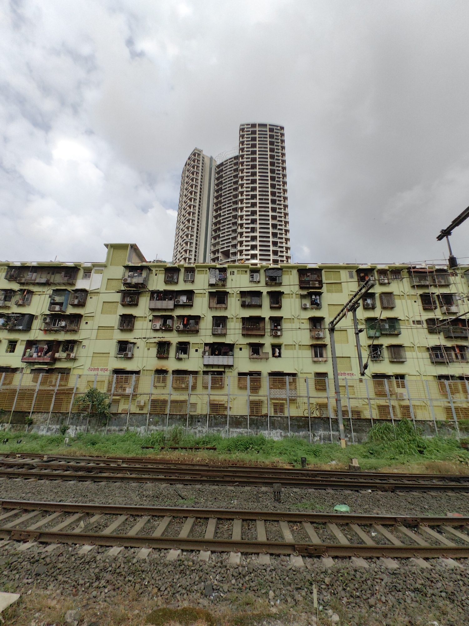 The image depicts a scene in Mumbai, India, showing a stark contrast between old and new architecture, situated adjacent to railway tracks. In the foreground, multiple railway tracks are visible, laid on a ballast of grey stones. To the left of the tracks, a patch of dry grass and some green vegetation is present.
Behind the tracks, a light yellow, multi-story building is prominently featured. This building appears older, with numerous windows and balconies, many of which are adorned with makeshift additions and air conditioning units. Some laundry is visible hanging from the balconies. A yellow metal fence runs along the front of this building, separating it from the railway line.
Behind the yellow building, two tall, modern skyscrapers with a beige exterior and a grid-like pattern of balconies rise into the overcast sky. The sky itself is filled with thick, grey clouds, suggesting a cloudy or potentially rainy day. Electrical wires and support structures are visible in front of the yellow building, suggesting it is located in a built-up urban area. A sign in Hindi is visible on the yellow building. A bicycle is leaning against the fence in front of the yellow building.