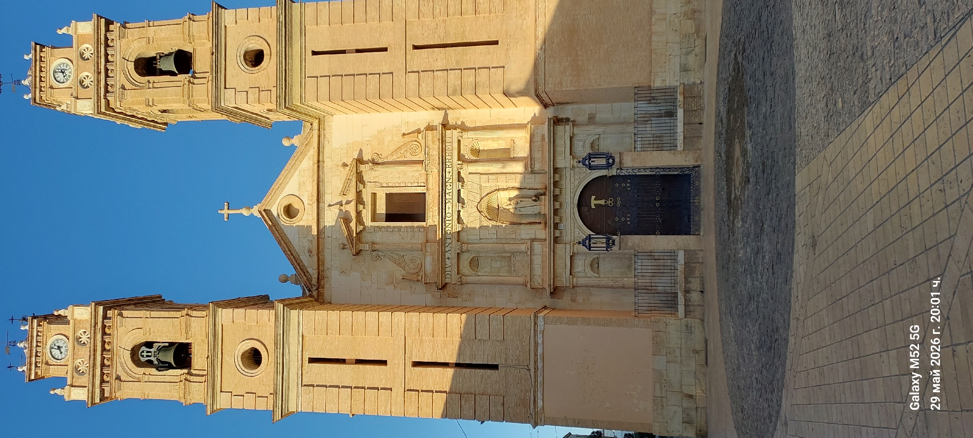 The image shows the façade of the church of San Antonio Abad in Canals, Spain. The church has two bell towers on either side of the main entrance. The towers are topped with clocks and weather vanes. The façade of the church is decorated with sculptures and carvings, including a statue of Saint Anthony of Padua. The main entrance has a large wooden door with a cross in the center. Two ornate lamps hang on either side of the door. The church is made of stone and is in a good state of repair. The sky is blue and clear. The image also contains the text "DIV. ANTONIO + MAG..." and the date and time of the photo being taken, which is May 29, 2026 at 20:01.