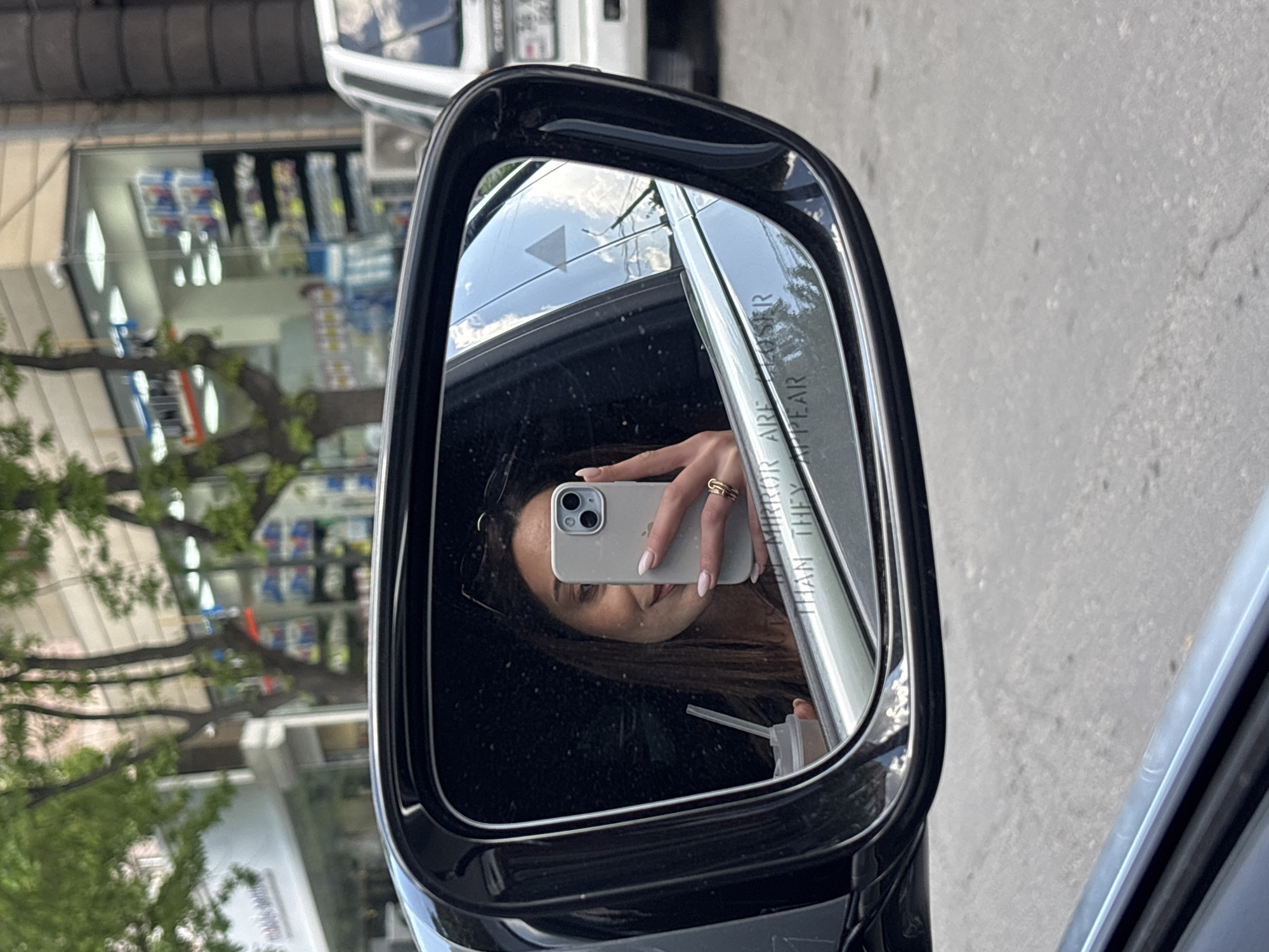A woman is reflected in a car's side mirror while holding an iPhone 14. She's wearing rings on her fingers, and a drink with a straw is visible. The mirror reads "OBJECTS IN MIRROR ARE CLOSER THAN THEY APPEAR". In the reflection, Yerevan's street scene is observable. A shop window filled with products can be seen, suggesting a commercial area.