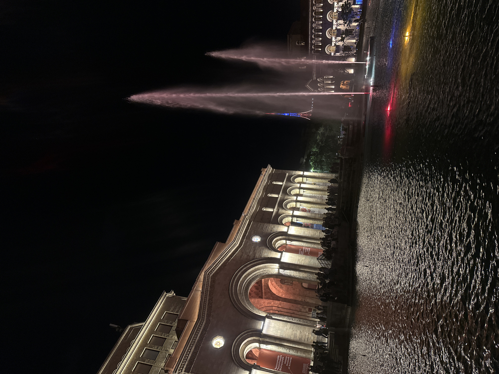 The image depicts a night scene of a fountain display in an urban setting, possibly in Unknown, Unknown. The lower portion of the frame features a building with illuminated arches and columns, exhibiting architectural details. People are seen on the balcony, suggesting an audience or spectators. In the background, water fountains are illuminated with colored lights, creating a visual spectacle against the dark night sky. The surface of the water reflects the surrounding lights, adding to the overall visual complexity.
