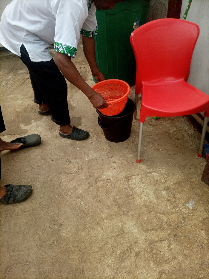 A person wearing a white shirt and black pants places an orange basket inside a black bucket. A red plastic chair stands to the right of the bucket. Behind the chair is a tall, green container. The person is wearing dark-colored sandals. Another person is visible in the lower left corner, also wearing dark-colored sandals. The ground is a textured, light brown color with imprinted designs.