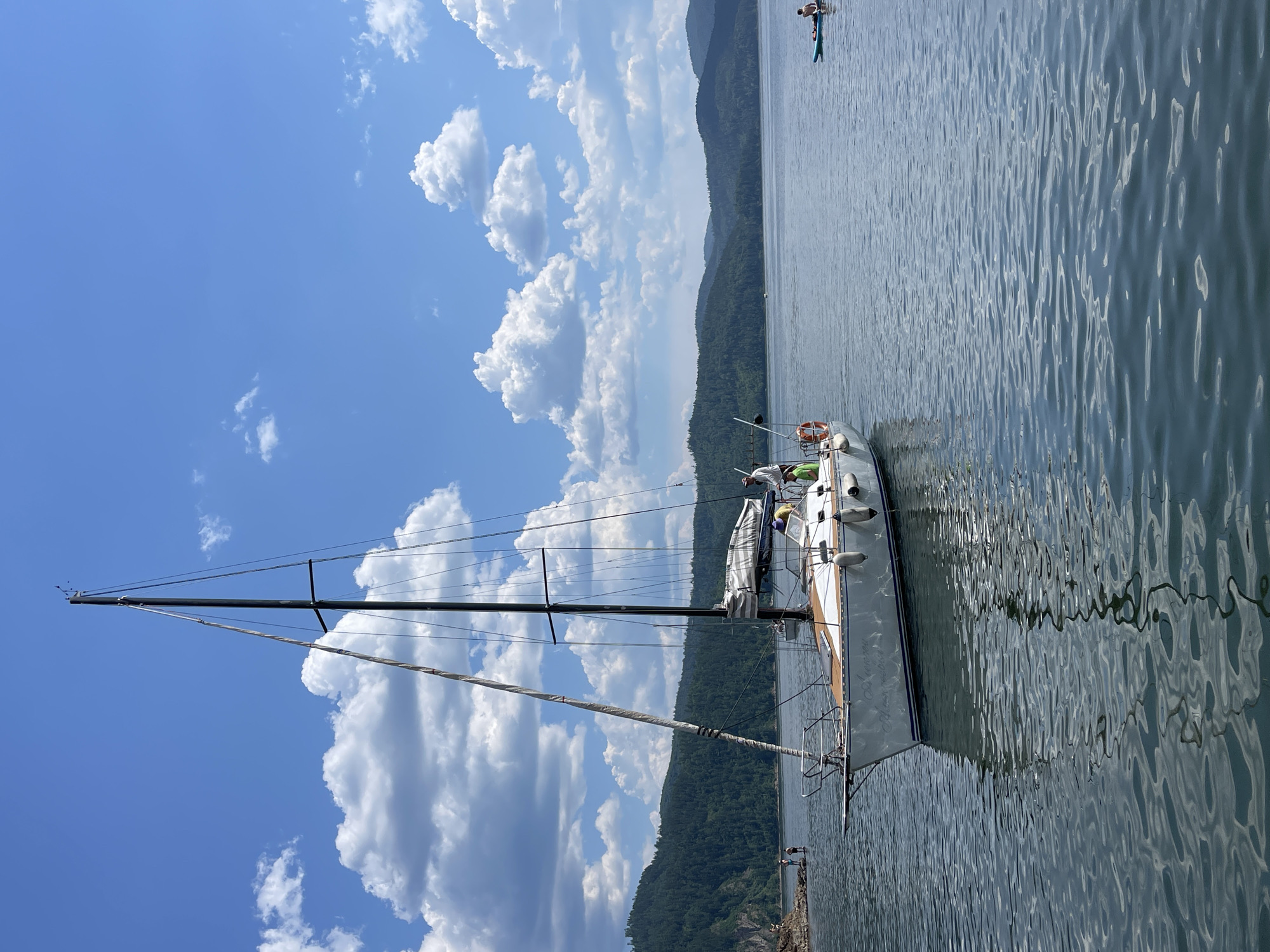 A sailboat is moored on a calm lake. The boat is white with wooden accents and has a tall mast. Two people are visible on the deck of the boat, and one person is on a paddleboard in the distance. The sky is blue with scattered white clouds. Lush green hills rise from the far side of the lake, suggesting a mountainous or hilly terrain. The water is slightly rippled, reflecting the sky and surrounding landscape. The lighting suggests it is daytime, likely late morning or afternoon, under clear, sunny weather. The scene evokes a sense of peaceful leisure and outdoor recreation on the water. The location is Divnogorsk, Russia. No visible text is present in the image.