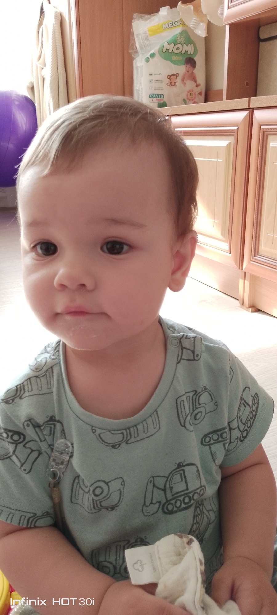 A young child is positioned in the foreground of the image, facing the camera. The child has light brown hair and is wearing a light blue t-shirt adorned with a pattern of construction vehicles. Visible on the child's chin are remnants of food or drink. The background features a shelf with a package of diapers, labeled "MEGA MOMI," and wooden cabinets. To the left of the frame, a portion of a purple exercise ball is visible. The lighting suggests an indoor setting, likely a home environment. The image is located in the city of Kedrovy, Russia.