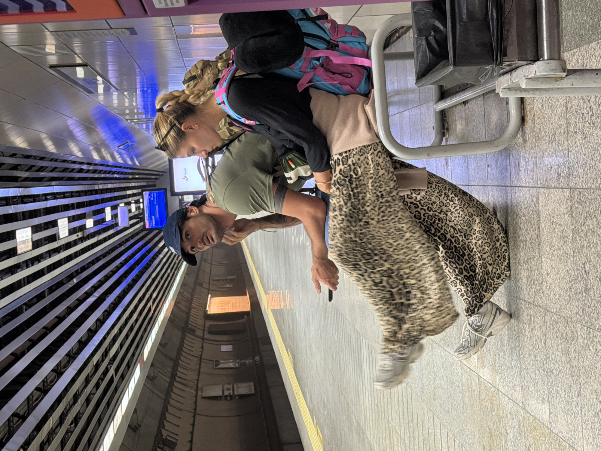 In Istanbul, Turkey, two individuals are seated on a metal bench at a subway platform. A man with a blue baseball cap, olive green t-shirt, and dark pants is looking to his left, with his left hand resting on his chin and holding a black smartphone. A woman with blonde hair in a braid, wearing a black long-sleeved shirt, a tan cardigan, and a leopard print maxi skirt, is looking to her right. She has a large backpack with blue and pink accents on her back. The subway platform has a yellow striped safety line along the edge and a polished concrete floor. The ceiling features a series of parallel metallic beams interspersed with lights. In the background, a dimly lit tunnel stretches into the distance, with some orange light emanating from an opening. A trash receptacle is visible to the right of the bench. The overall scene depicts a moment of waiting on a public transportation platform.