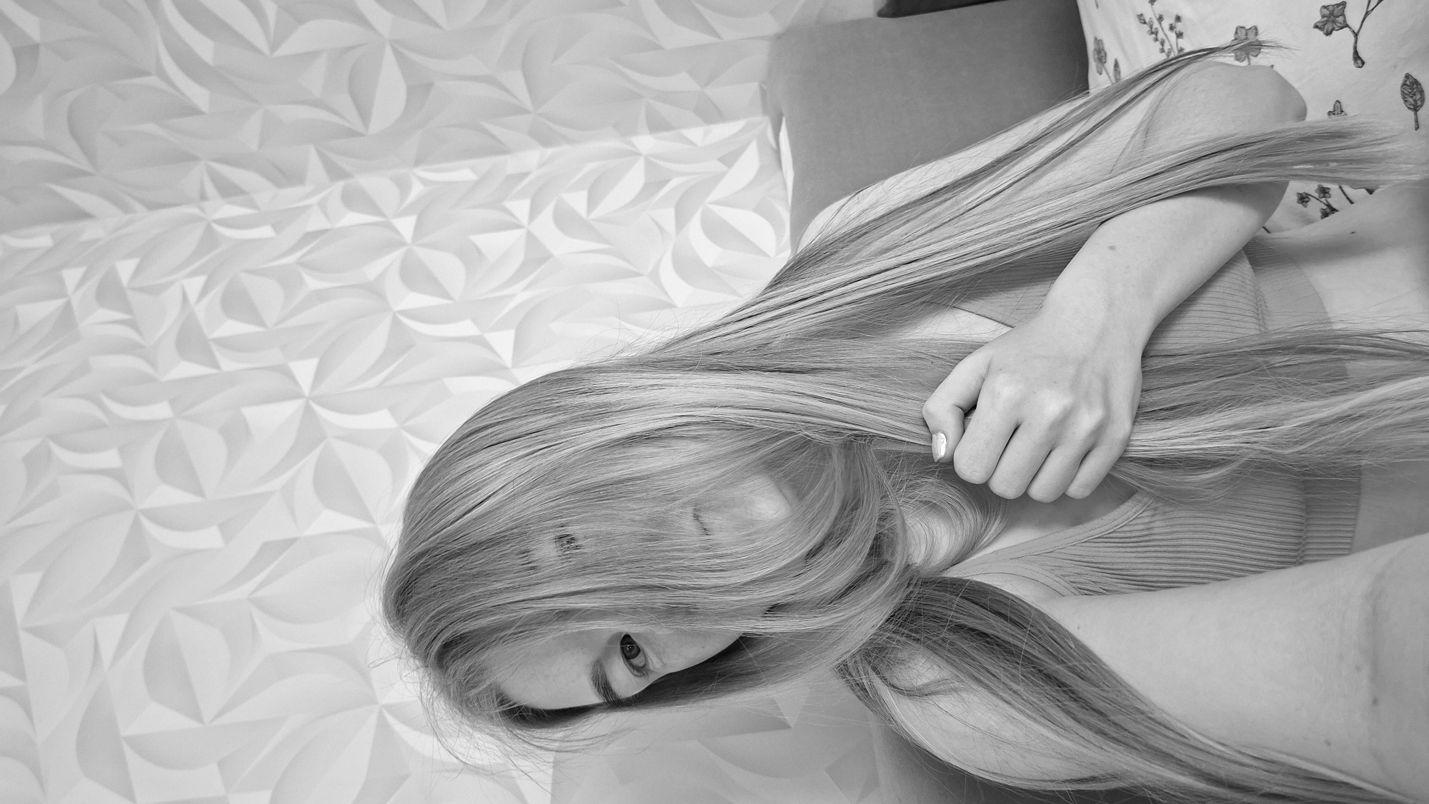 A black and white, high-angle shot shows a person with long hair and a hand with painted nails. The person is lying with their head down, and their hair is spread out. A hand is visible, holding the hair. A patterned wall or surface is visible on the left side of the image. The location in the city of Chaikovsky, Russia, cannot be determined from the image.
