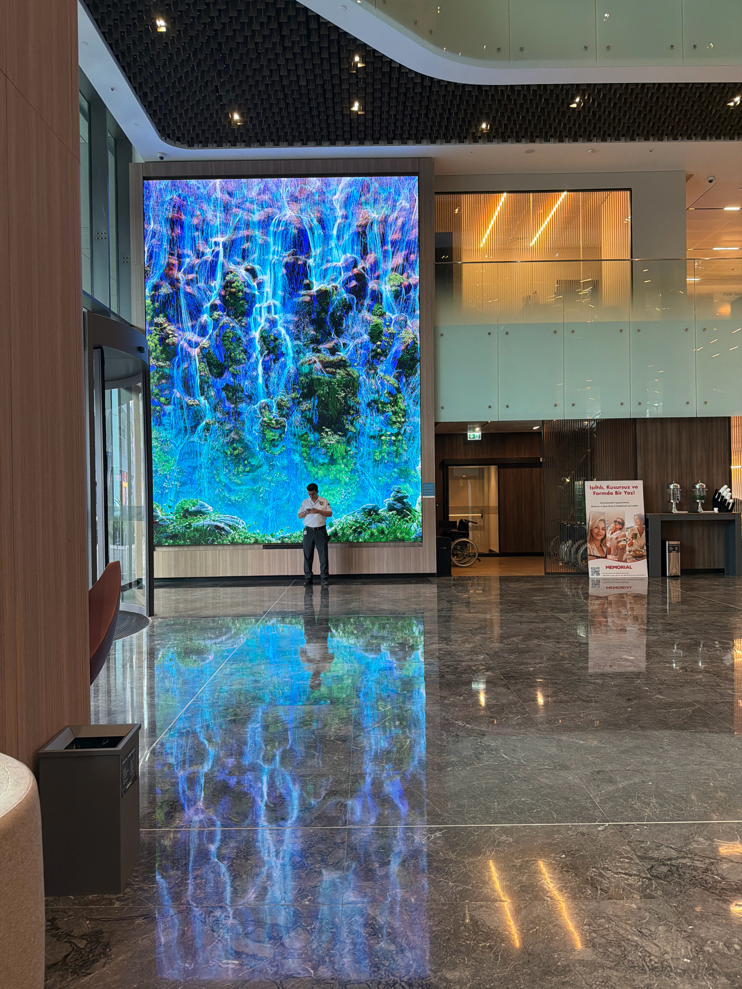 A man stands in front of a large digital display showing a vibrant, blue waterfall, reflecting on the polished marble floor in a modern building lobby. The man is wearing a white collared shirt and dark pants and is looking at his phone. To the left, a revolving glass door leads outside. To the right, a promotional poster for Memorial Healthcare Group is displayed, featuring images of smiling people and text in Turkish. Further back, a wheelchair is visible near an open doorway, and a table with drinks and cups is set up. The ceiling features a patterned design with recessed lighting. The scene appears to be in the lobby of a commercial building, likely a hospital or healthcare facility, in Ataşehir, Turkey.