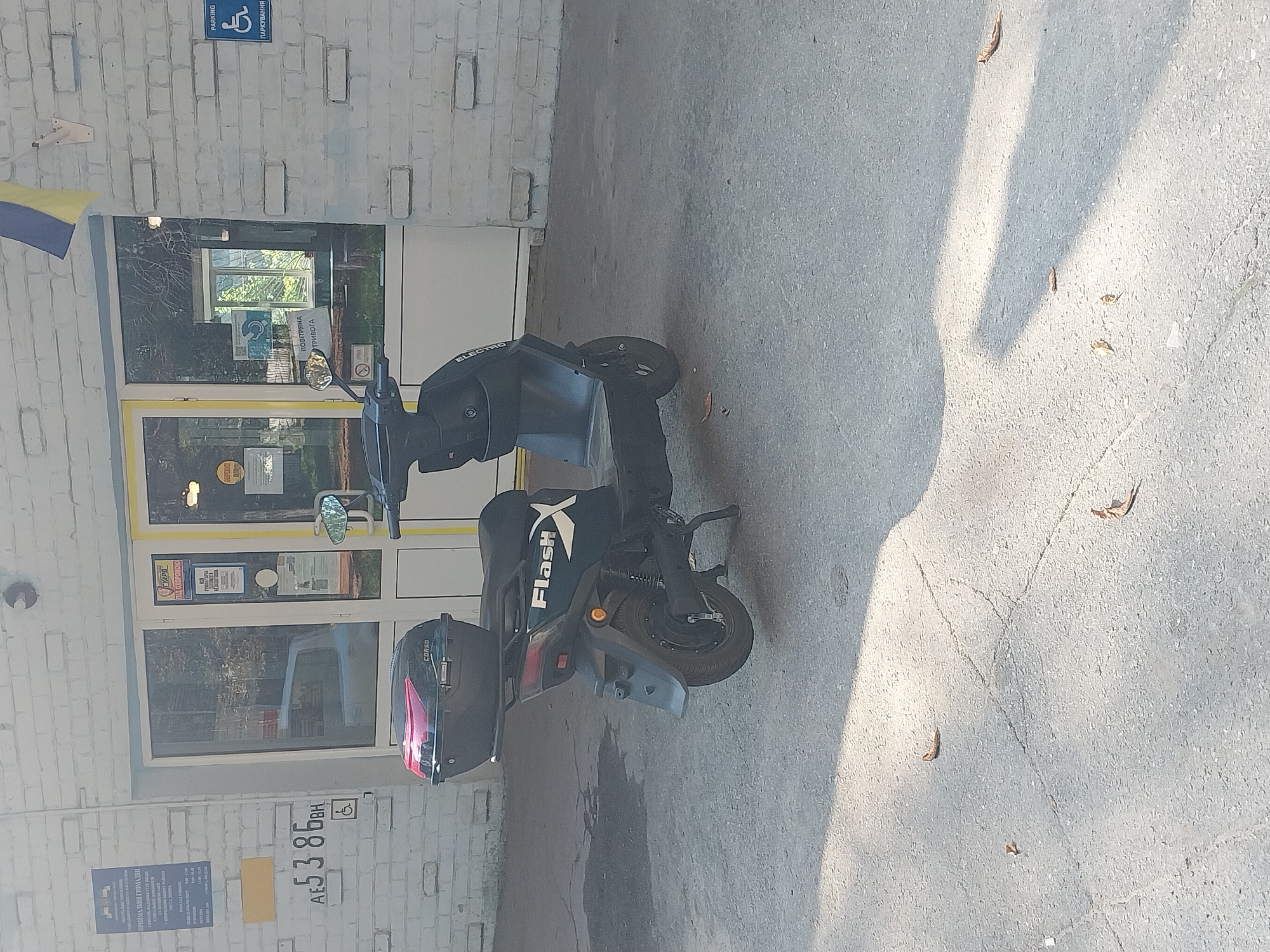The image shows a black electric scooter parked on a paved surface next to a brick building. The scooter has "FlashX" and "ELECTRO" written on it, and a black helmet with red accents is attached to the back. In the background, there is a building with large windows, some of which display signs. One sign appears to be a "Parking" sign with a wheelchair symbol, and another reads "POвітряна ТРИВОГА" (Air Raid Alert) in Ukrainian. There is also a Ukrainian flag visible on the left side of the building. The lighting suggests it is daytime, possibly late afternoon, with shadows cast on the ground. The scene appears to be an urban or semi-urban environment. The overall impression is one of everyday life potentially impacted by current events, given the presence of the air raid alert sign.