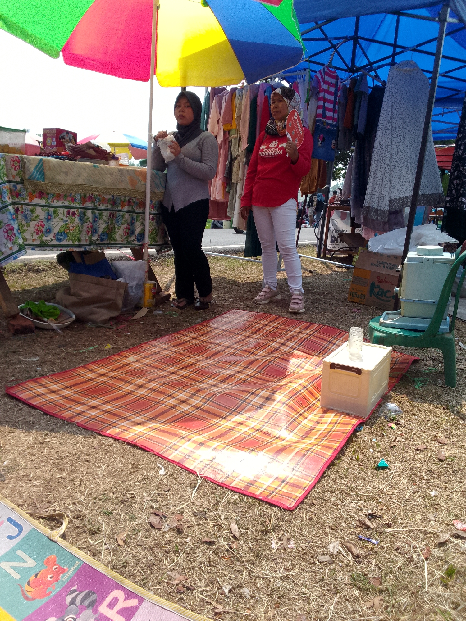 This is a medium-shot, eye-level, outdoor photograph taken in Mandalawangi, Indonesia, depicting a street market scene. Two women are standing under colorful umbrellas and a blue tarp that provides shade. One woman, wearing a grey hijab and long-sleeved grey shirt with black pants, stands behind a table covered with floral fabric. She is holding a white plastic bag or wrapper in her right hand and appears to be eating. The other woman, wearing a patterned headscarf and a red long-sleeved shirt with white pants, stands further back. She is holding a red fan in her left hand and looking towards the viewer.
In the foreground, a red and orange plaid mat is laid out on dry, brown grass and scattered leaves. To the right of the mat, a white plastic cooler box with a glass of water on top sits next to a green plastic chair. Behind the women, clothing is displayed on hangers, and various items, including boxes, bags, and a yellow cylindrical object, are visible under the market stall. The ground is covered with dry grass, dirt, and small debris. In the lower left corner, a portion of a colorful mat with children's alphabet illustrations is visible. The lighting suggests it is daytime, likely sunny, with some shadows cast.