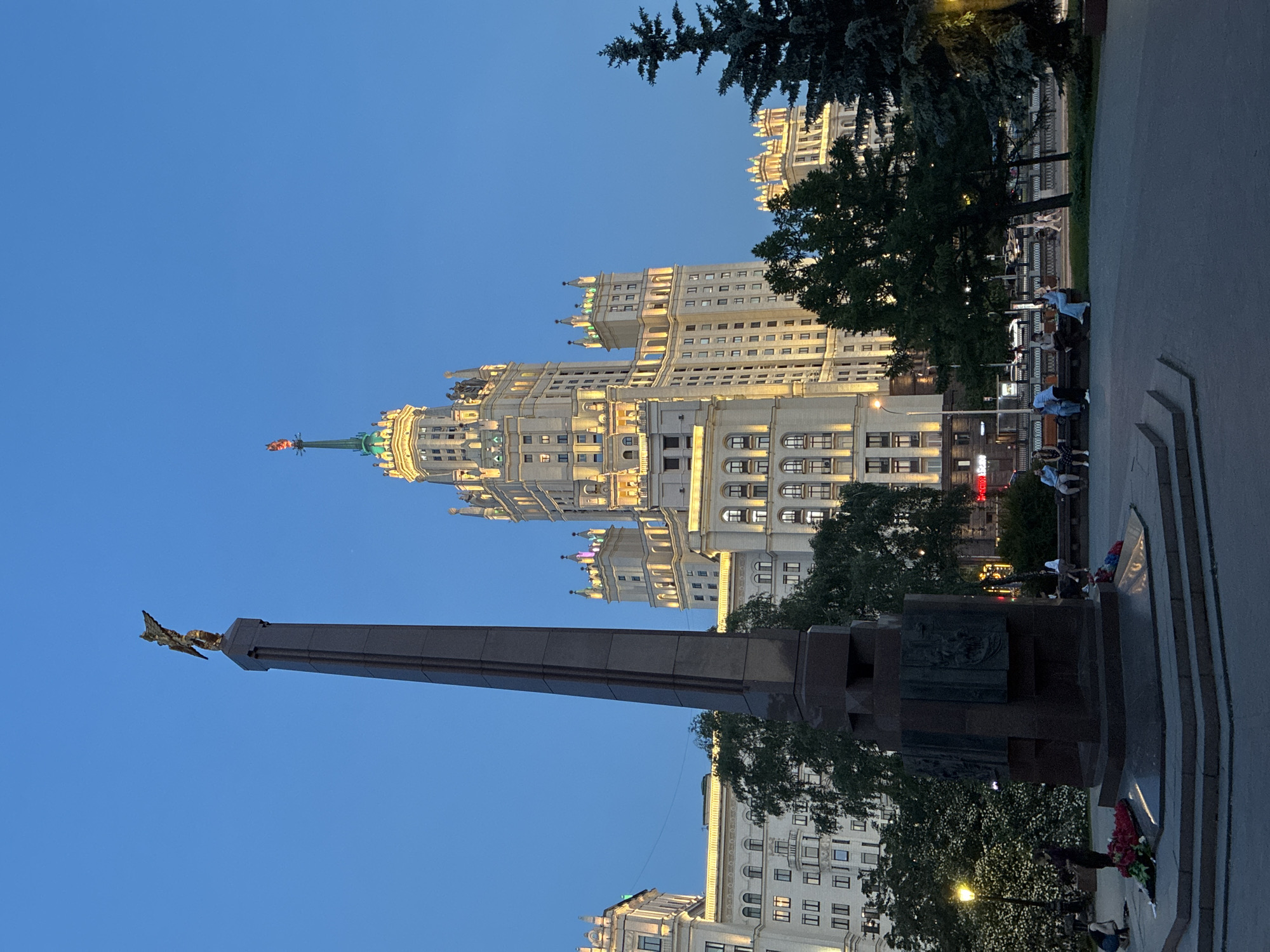 The image captures a view in Moscow, Russia, featuring the Kotelnicheskaya Embankment Building, one of the Seven Sisters skyscrapers. In the foreground is a monument topped with a golden eagle. People are seen sitting on benches near the monument. The sky is a clear blue. The buildings are illuminated with artificial light. Trees are present near the monument.
