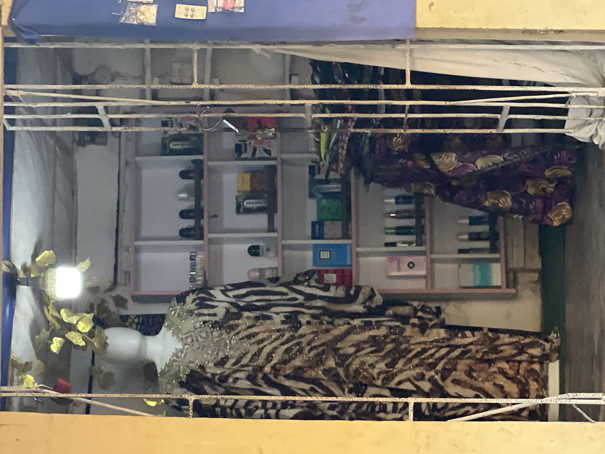 The image shows the interior of a small storefront, likely a boutique, viewed through a barred entrance. The storefront is located in Nkpor, Nigeria. A mannequin dressed in a long, flowing garment with a zebra print pattern and elaborate beaded embellishments on the neckline stands prominently on the left. Behind the mannequin, white shelves are visible, stocked with various items, including what appear to be bottles of perfume or cosmetics, and some boxed products. The shelves are arranged in a staggered pattern, with some items stacked higher than others. The lighting in the store is dim, with a single bright light fixture hanging from the ceiling above the mannequin, illuminating some artificial yellow leaves draped nearby. On the right side of the image, more fabric, possibly more clothing or textiles, is visible, stacked or draped. The entrance is secured by a metal grate, with one of the bars showing rust. A blue tarp or fabric is visible on the far right, partially obscuring the view of the shop's interior. The overall impression is of a retail space displaying clothing and other merchandise, with a somewhat cluttered but functional arrangement.