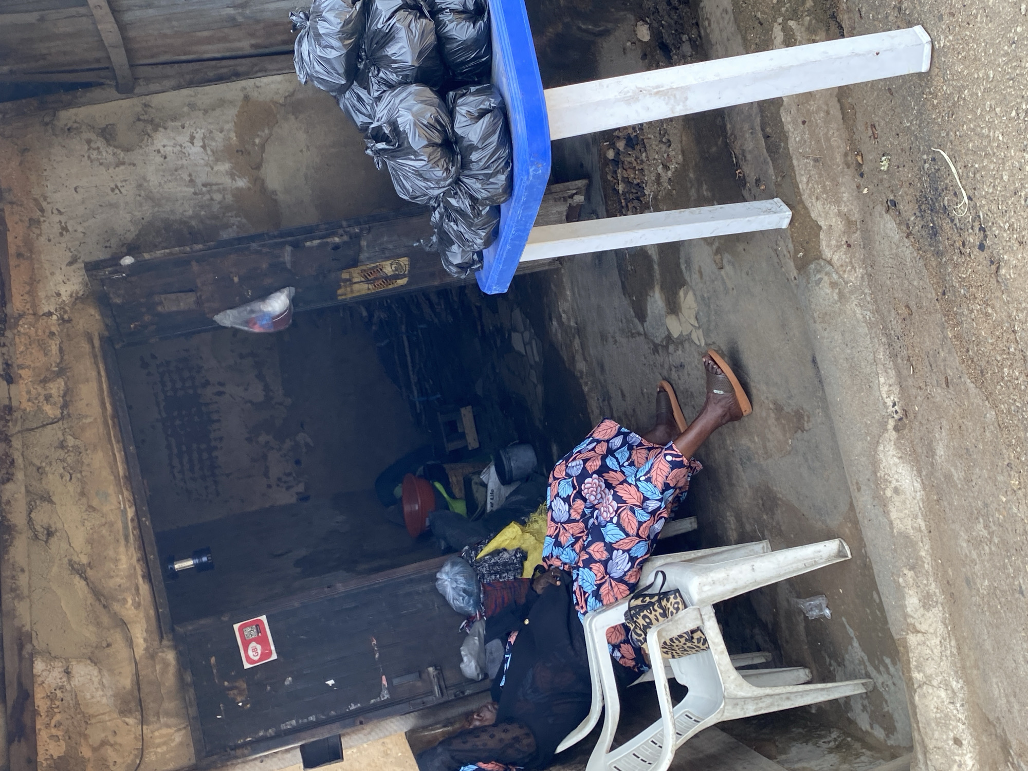 In Kabong, Nigeria, an elderly woman wearing a black headwrap and a blue and pink floral dress is seated in a white plastic chair, appearing to be asleep. A leopard-print purse rests on the chair beside her. To her right, a blue table holds a stack of black plastic bags filled with unknown contents. Behind the woman, a dimly lit doorway leads into a store, where various items, including a red bowl and more bags, are visible. The ground in front of the store is dirt and concrete, with some debris scattered around. The overall environment suggests a busy marketplace or street-side stall.