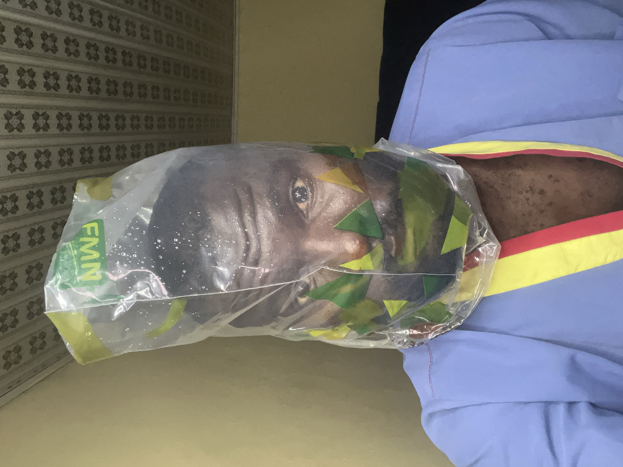 A dark-skinned man's head is encased in a clear plastic bag. The bag features some green and yellow triangular shapes. The man's face is visible through the bag and beads of water are condensed on the inside of the bag. The man is wearing a blue and yellow jacket. The background features a wall with an ornate pattern of geometric shapes and another light-colored wall. The location is Gura Top, Nigeria.