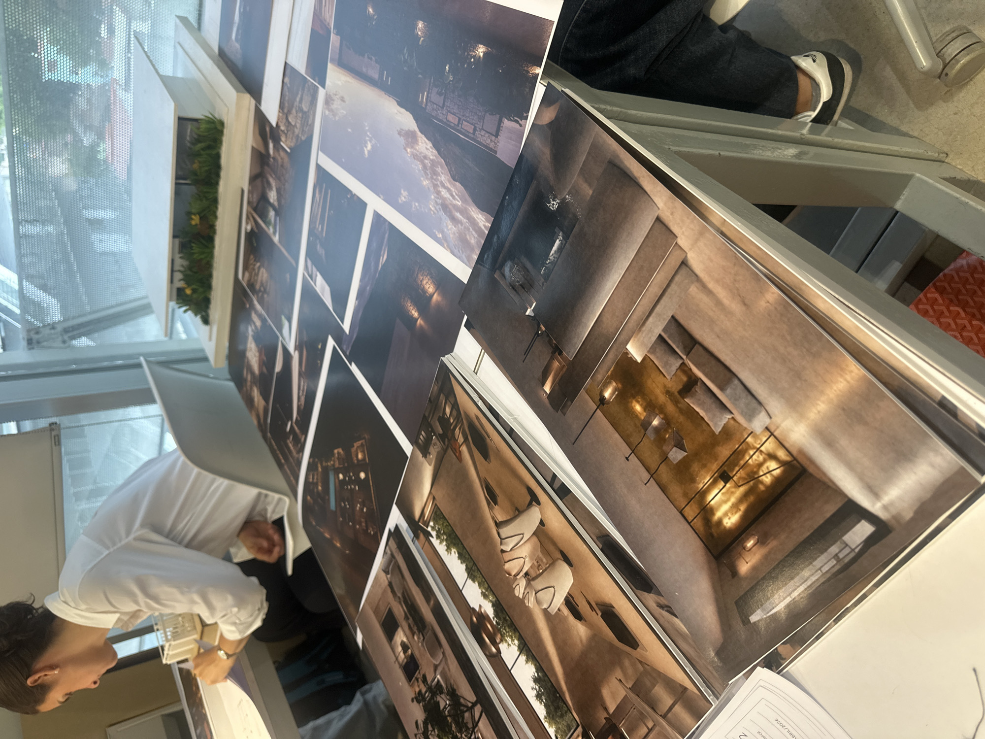 The scene shows a person examining architectural renderings or prints displayed on a table, potentially in Mexico City, Mexico. The renderings feature modern interior and exterior designs with an emphasis on lighting and spatial arrangements. The person appears to be dressed in a white shirt. The environment includes a building's interior, possibly a studio or office setting, with plants in a window box. In the background, someone is sitting in a chair.