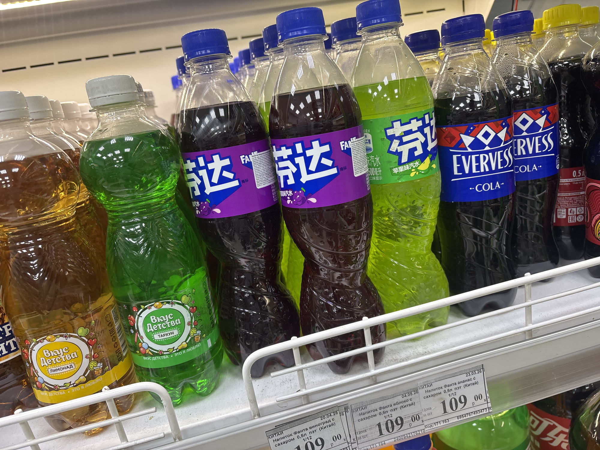 The image shows a shelf stocked with various flavored sodas in a store, possibly in an Asian city, judging by the Chinese text on the Fanta bottles. The sodas include "Вкус Детства" (Taste of Childhood) lemonade and tarragon flavors, Fanta in grape, apple, and pineapple flavors, and Evervess Cola. All the sodas are in clear plastic bottles with labels. The Fanta bottles have blue caps, while the "Вкус Детства" bottles have white caps. The prices of the Fanta sodas are displayed as 109.00 on printed labels below the bottles.