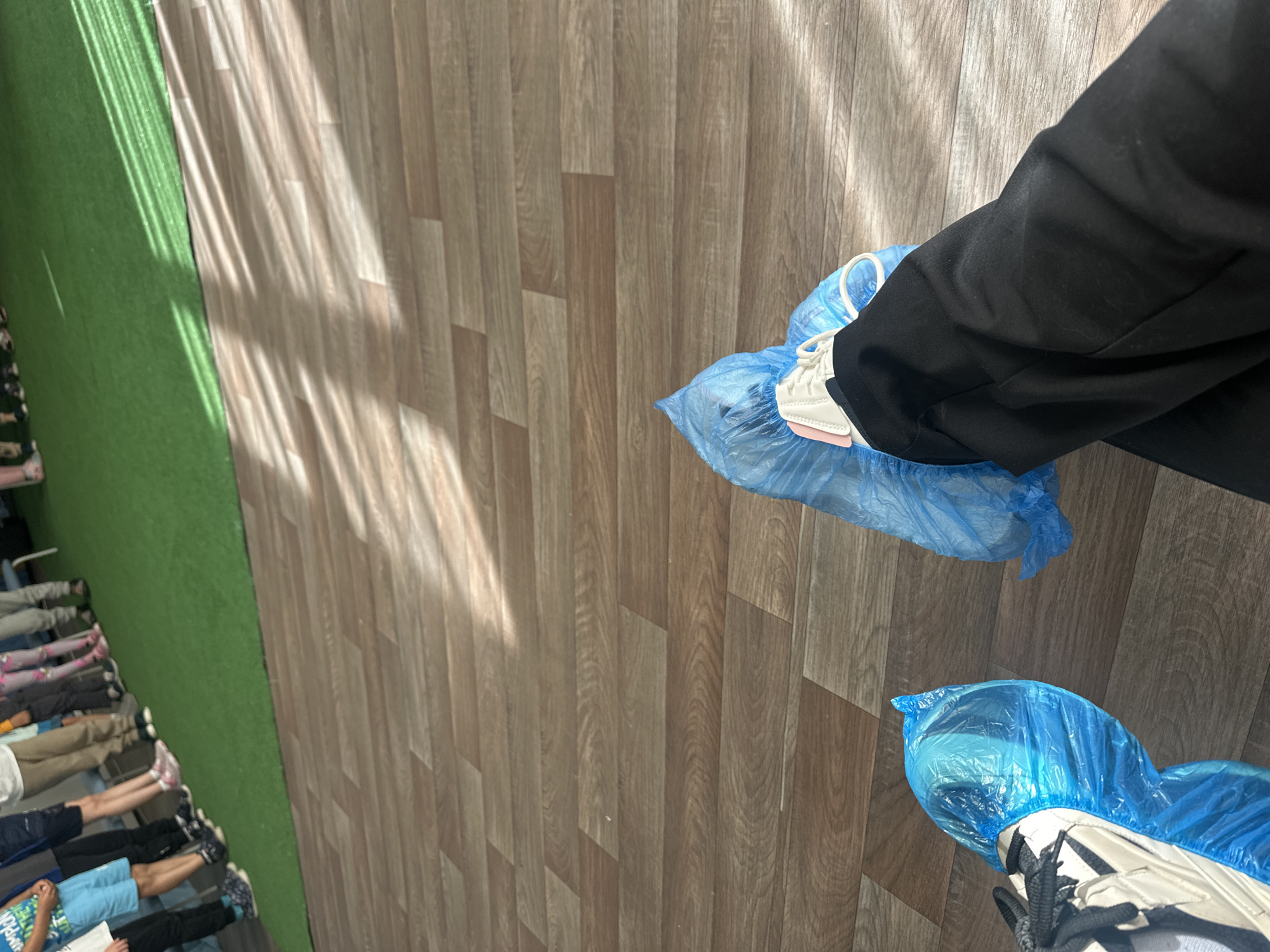 Two individuals are wearing blue plastic shoe covers over their footwear while standing on a wood-patterned floor. One person is wearing black pants. In the background, there's a strip of green turf and a row of people sitting in chairs. The location is possibly in Plovdiv, Bulgaria.