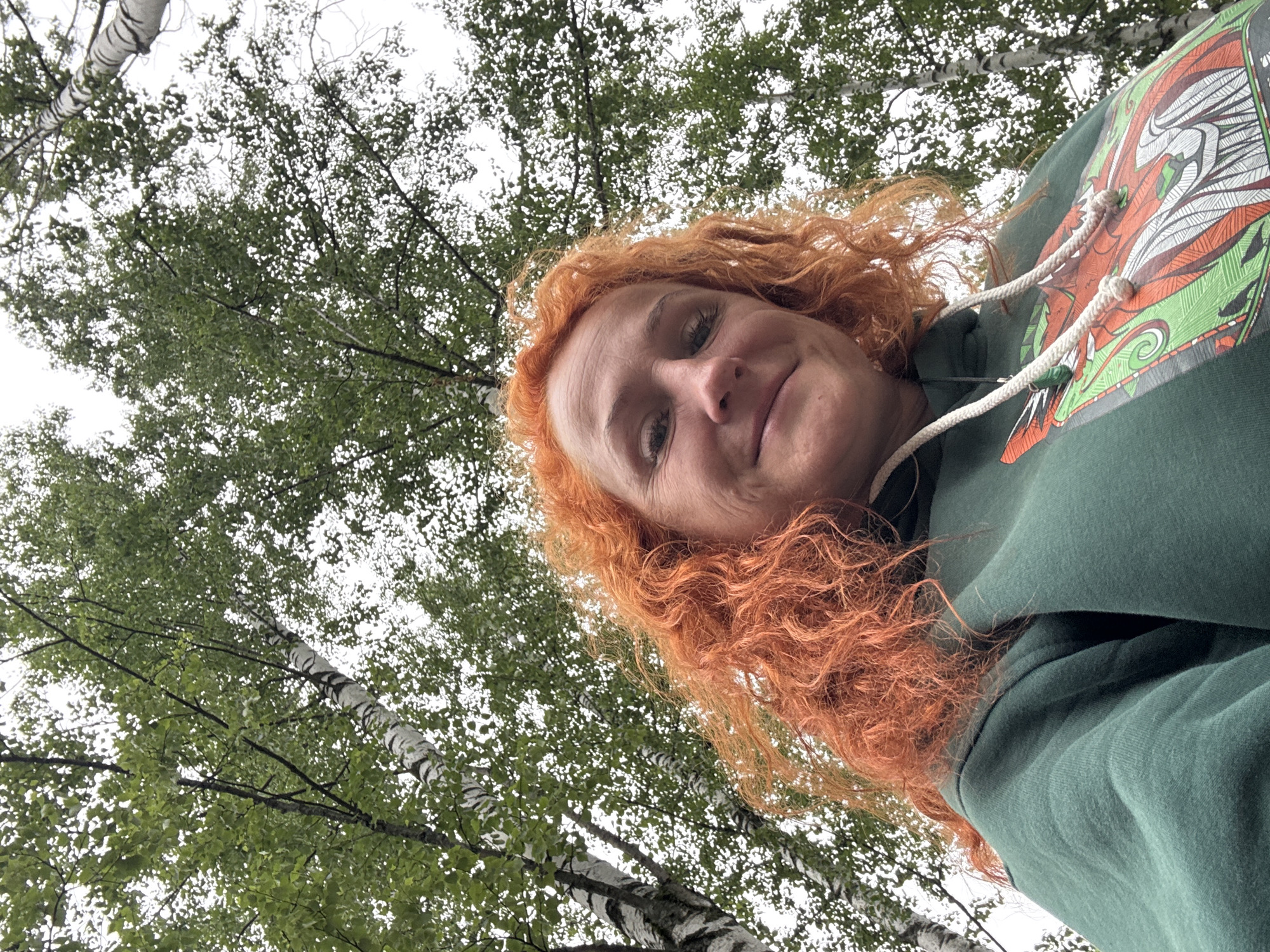 A low-angle, close-up selfie shows a woman with bright orange, curly hair, smiling slightly and looking upwards. She is wearing a dark green hooded sweatshirt with a graphic design of a dragon-like creature in orange and green. The background is dominated by the canopy of birch trees, with their distinctive white and black striped bark visible. The sky is overcast and white. The scene appears to be outdoors in a park or forest area. It is not possible to determine the specific location within Chaikovsky, Russia from the image alone.