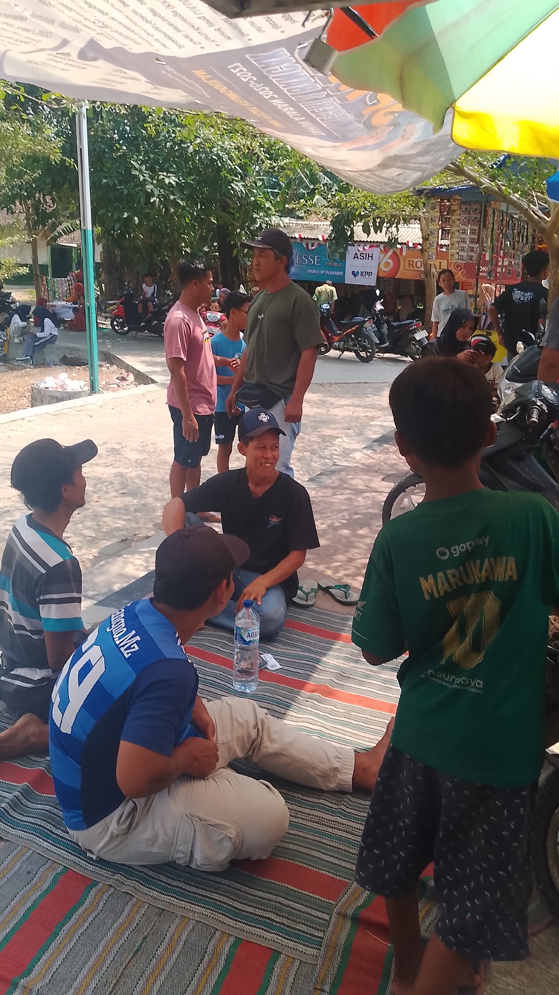 In Karanglangu, Indonesia, under a canopy, several people are gathered outdoors in a casual setting. Three young men are seated on a striped mat on the ground. One man, wearing a blue and black striped t-shirt and a dark baseball cap, is on the left, facing away from the camera. Next to him, a man in a blue jersey with the number "14" is seated, also facing away from the camera. In the center, a man in a black t-shirt and blue jeans is sitting cross-legged, looking towards the left and smiling. A bottle of Aqua water is on the mat in front of him. To the right of the seated individuals, a young boy in a green t-shirt with "MARUKAWA" printed on it is standing, facing away from the camera.
In the background, several other people are visible. Two men stand near a white tent structure displaying various advertisements. One man is wearing a pink t-shirt and dark shorts, and the other is in a green t-shirt and blue jeans. Behind them, a row of stalls with goods displayed suggests a marketplace or community gathering. Motorcycles are parked in the area, indicating a mode of transportation. The environment appears to be outdoors, with trees and sunlight filtering through the canopy. The ground is covered in a mixture of sand and paved areas.