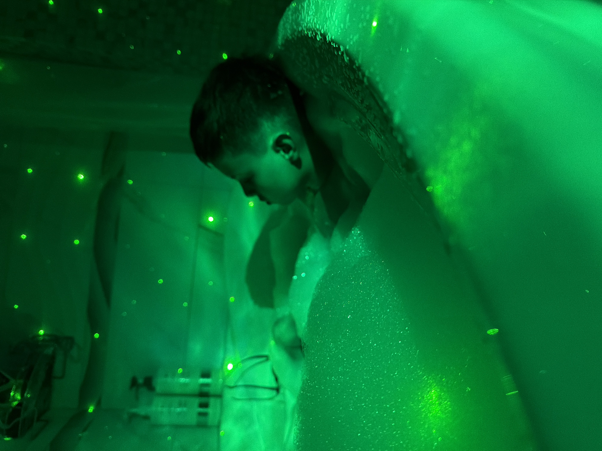 A young child is in a bathtub filled with bubbles, illuminated by green lights, including small points of light across the ceiling. The location in Votkinsk, Russia is not discernible from the image. The child is looking down, partially obscured by the bubbles. There are objects on a shelf or ledge beside the tub, including a small fan, bottles and cords.