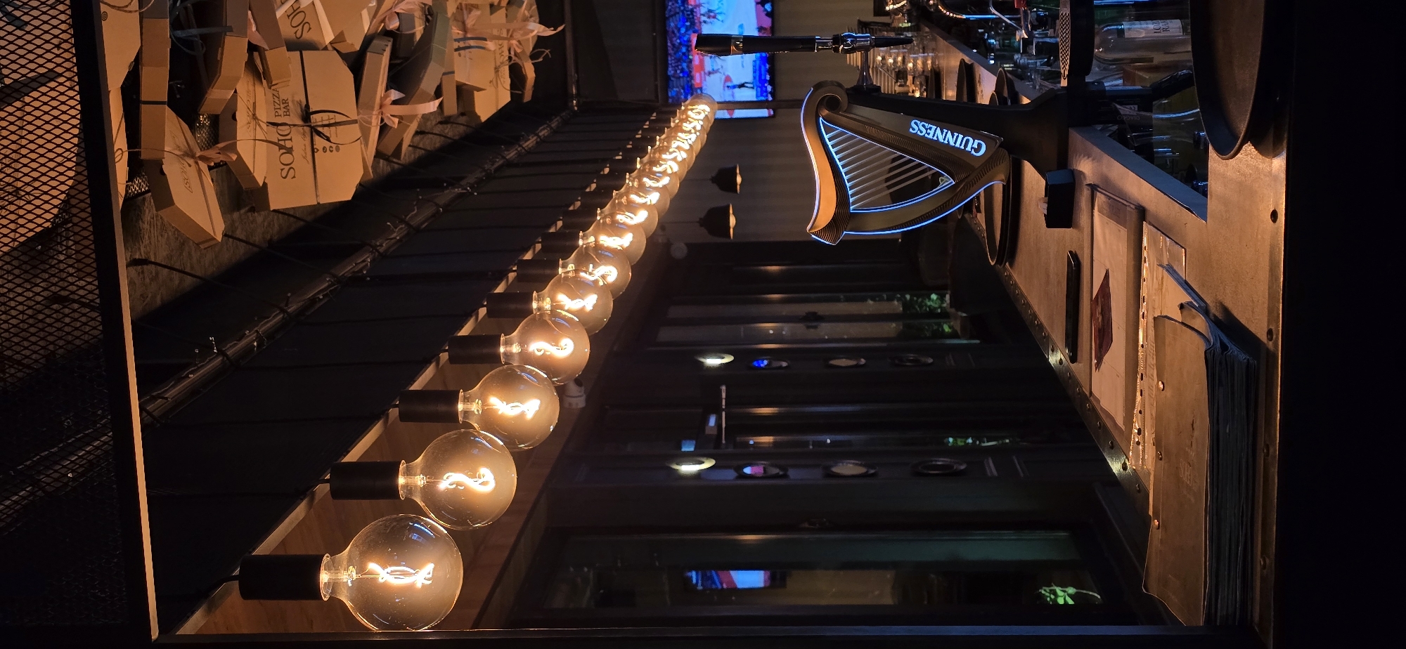 The image shows the interior of a bar, likely in Bar, Montenegro, based on the pizza boxes that have the name "SOHO" written on them, as well as other signage. A row of lit, vintage-style light bulbs extends into the background. In the right of the frame is a Guinness sign, and a counter area with menus and what appears to be a metal edging. Above, various bottles are visible. In the background is a TV screen.