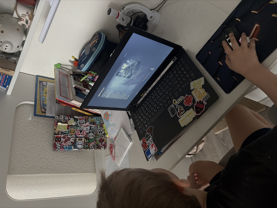 A young individual, viewed from behind, is seated at a white desk in what appears to be a study area within a residence in Novosaratovka, Russia. The person, with light-colored hair and wearing a dark t-shirt and shorts, is operating a black laptop computer. Their right hand is positioned on a white and black gaming mouse, which rests on a dark gaming mousepad with orange accents.
The laptop's screen displays a menu labeled "ВЫБОР МИССИИ" (MISSION SELECT) with an image of what appears to be a snowy, war-torn environment, indicating a video game is active. The laptop's palm rest and surrounding surfaces are extensively covered with various stickers, including designs related to "NBA," "Android," "Star Wars," and text such as "GIRLFRIEND" and "GOT MY WIFI FLY FOR EDI."
On the left side of the desk, next to the person, are several scattered papers and notebooks, including a spiral-bound notebook with handwritten entries and a red pen. Further left, a tablet or similar device encased in a heavily stickered cover stands upright. On the right side of the desk, behind the laptop, is a blue race car-shaped pencil case. A white microscope with red detailing is positioned on the far right.
Above the desk, a white shelf is visible, holding a white piggy bank with dark spots, a small rocking horse figurine, and an assortment of books and papers, including one featuring Russian text. The immediate background shows a light-colored wall or furniture, possibly a cabinet. The floor under the desk is light-colored.