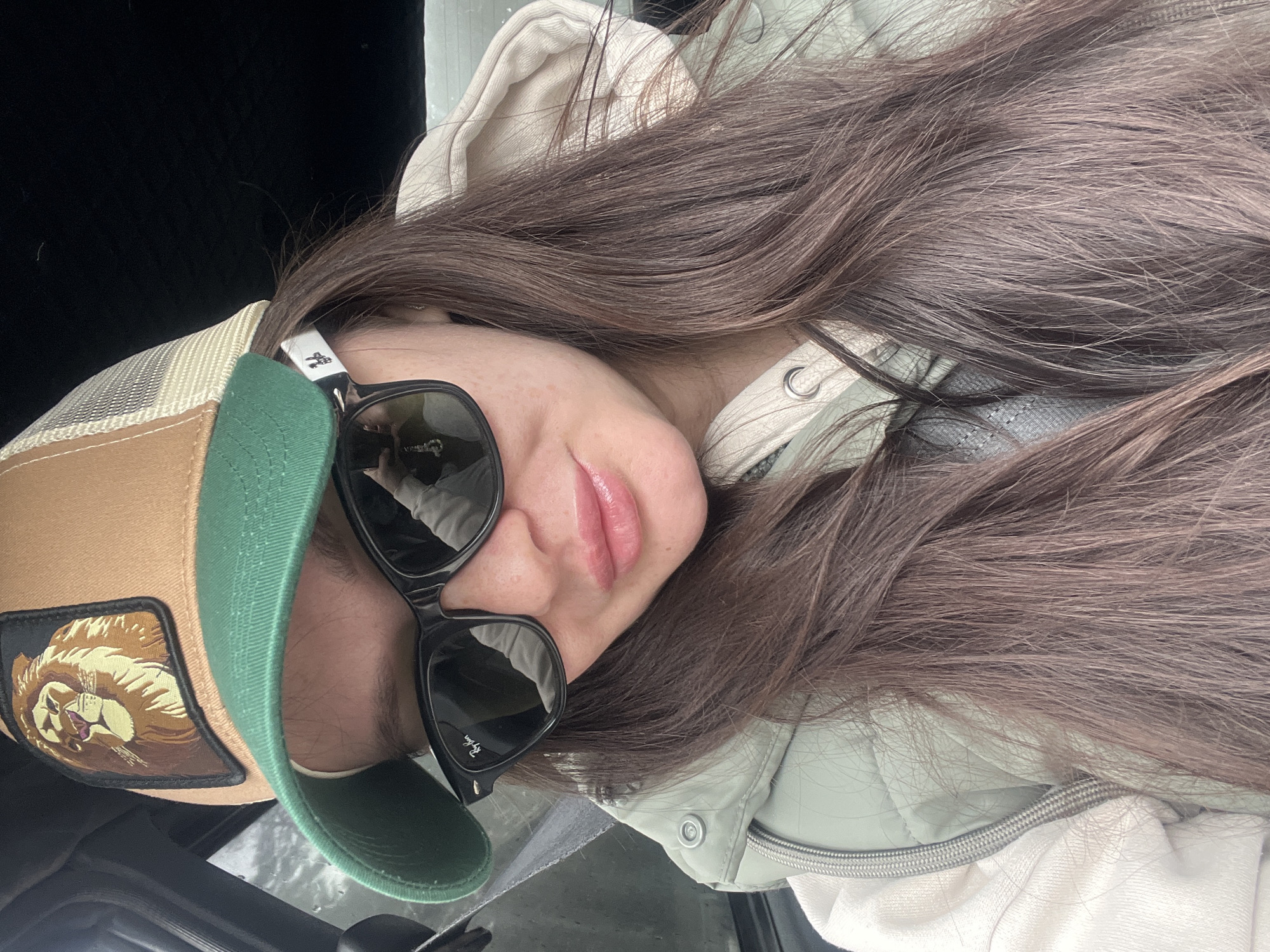 A woman is pictured in an indoor setting, likely a car. She is wearing a tan and green baseball cap with a lion patch, black sunglasses, and a light green quilted jacket with a white hooded sweatshirt underneath. The woman has long, brown hair. The black interior of the car is visible in the background.