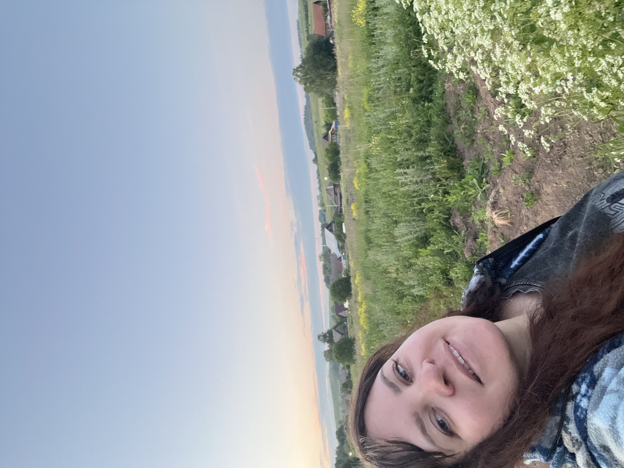 A selfie captures a woman in the foreground, with her face tilted towards the camera. She has long, dark brown hair and appears to be smiling. She is wearing a dark grey t-shirt and a patterned jacket.
In the background, a vast green field stretches out, dotted with yellow wildflowers and interspersed with small plants. Beyond the field, a village is visible, characterized by various houses with different roof colors, surrounded by trees. The sky above is a pale blue, with soft clouds tinged with pink and orange, suggesting it is either dawn or dusk. The overall scene depicts a rural landscape in Panteleevka, Russia.