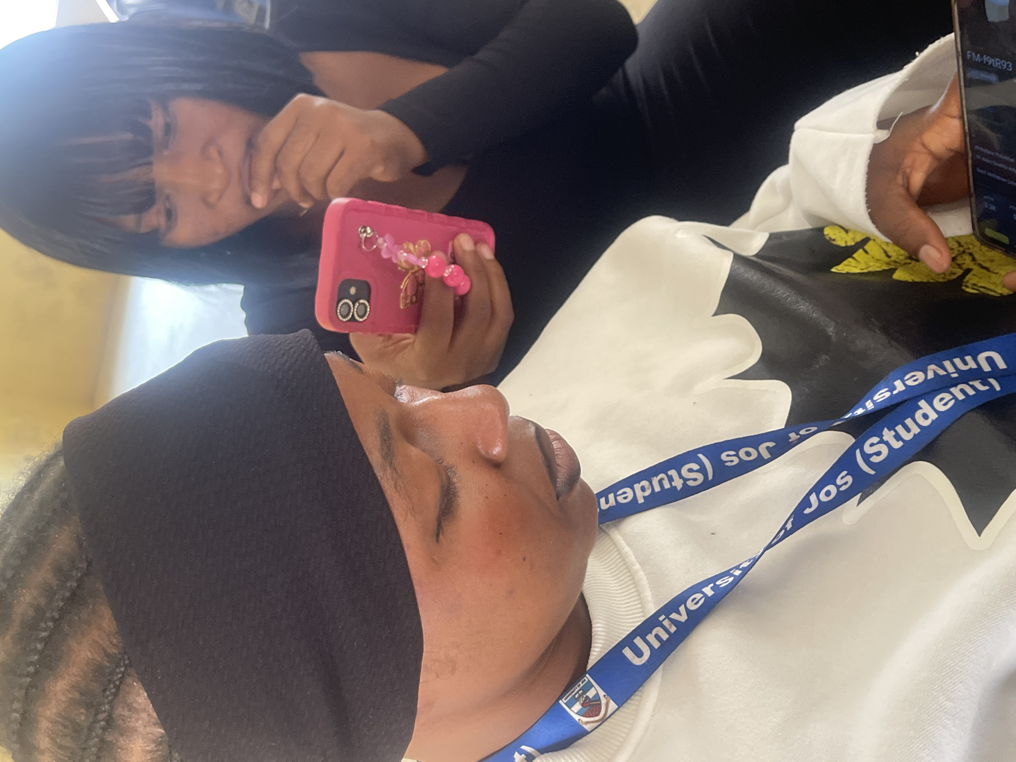 Two women are seated close together. The woman in the foreground, on the left, is wearing a black headband and a white sweatshirt with a black and yellow graphic design. A blue lanyard with "University of Jos (Student)" printed on it is around her neck. She is looking down at a smartphone. The woman in the background, on the right, has dark hair with bangs and is wearing a black top. She is holding a pink smartphone with a beaded charm and appears to be touching her face. The environment appears to be indoors, with a light-colored wall and ceiling visible. There is no specific indication of the location being Rafin Par, Nigeria, but based on the lanyard, it is likely the University of Jos.