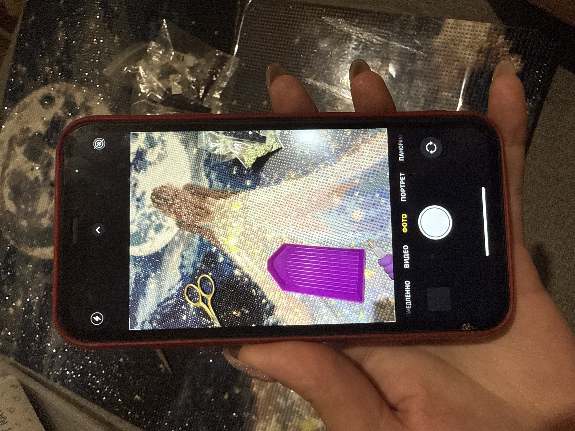 A person is holding a red iPhone in their hand, taking a photo of a diamond painting in progress. The painting appears to depict a celestial scene with a figure, possibly an angel or a woman, with flowing hair. There is a moon in the background. A purple diamond tray and a pair of gold scissors are visible on the painting surface. The image may have been taken in Dmitrov, Russia, though this cannot be confirmed from the image alone. No specific location within Dmitrov can be determined from the image.