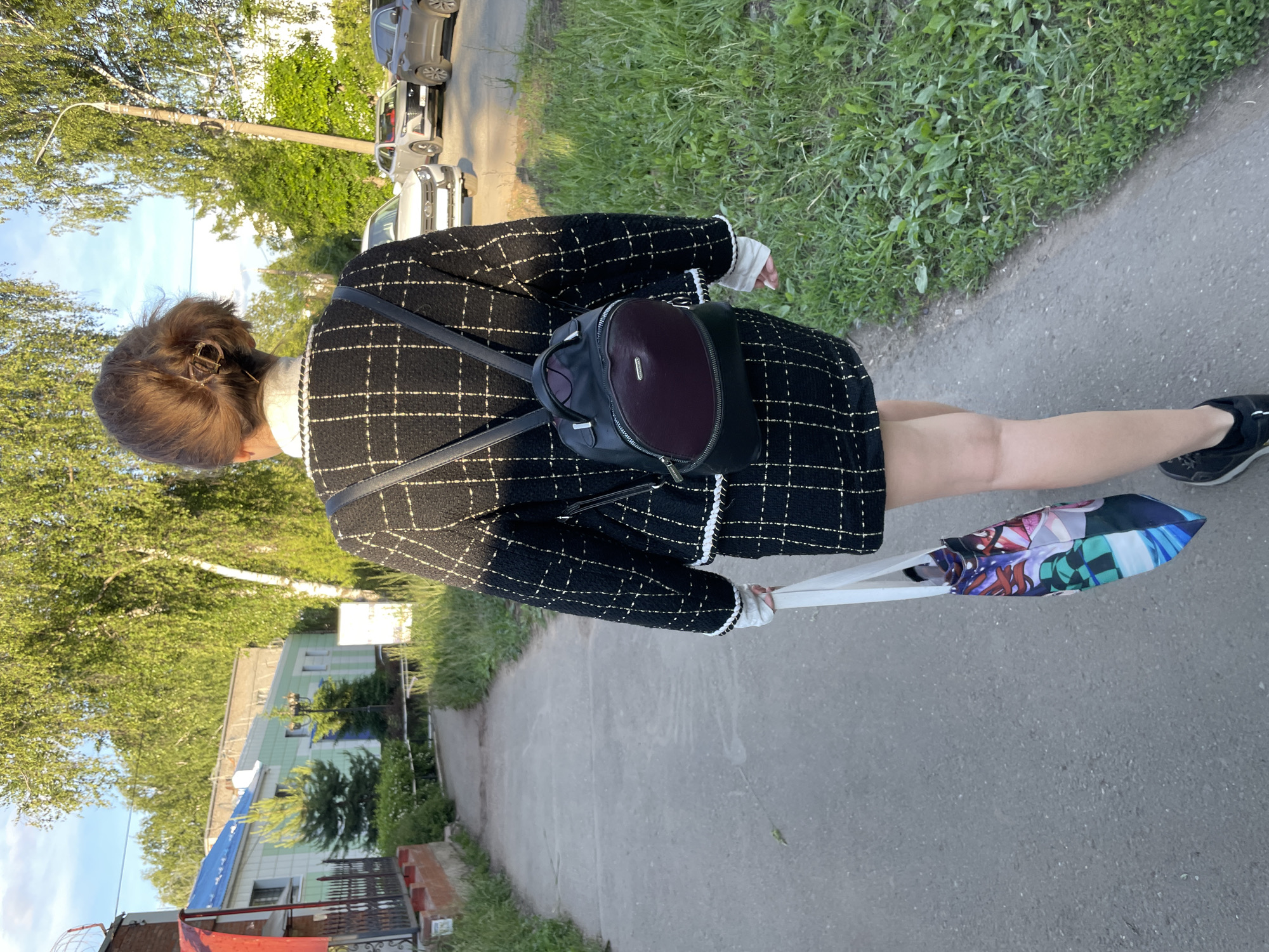 A person is seen from a rear, slightly elevated perspective, walking outdoors on a paved path. The subject has short, dark brown hair secured with a golden-colored hair clip. They are wearing a black and white checkered jacket or coat with white trim, a light-colored top with a high collar, and black athletic shoes. A dark-colored backpack, appearing to be black with a maroon section, is worn on their back. The person is also holding a folded umbrella with a colorful, anime-style pattern, including what resembles the green and black checkered pattern of Tanjiro Kamado's haori from Demon Slayer.
The setting appears to be a suburban or semi-urban street in Izhevsk, Russia. To the person's left, there is a grassy area leading to trees with green foliage under a clear blue sky with some scattered clouds. A street lamp post is visible further in the background. To the right of the person, there is a grassy verge, beyond which are parked cars on what looks like a dirt or gravel shoulder. Further in the background, residential or public buildings are visible, including one light green and white building with a blue roof and a metal fence in front. The light suggests it is daytime, possibly morning or late afternoon. The activity depicted is a casual walk. No specific readable text is visible on signs or objects.