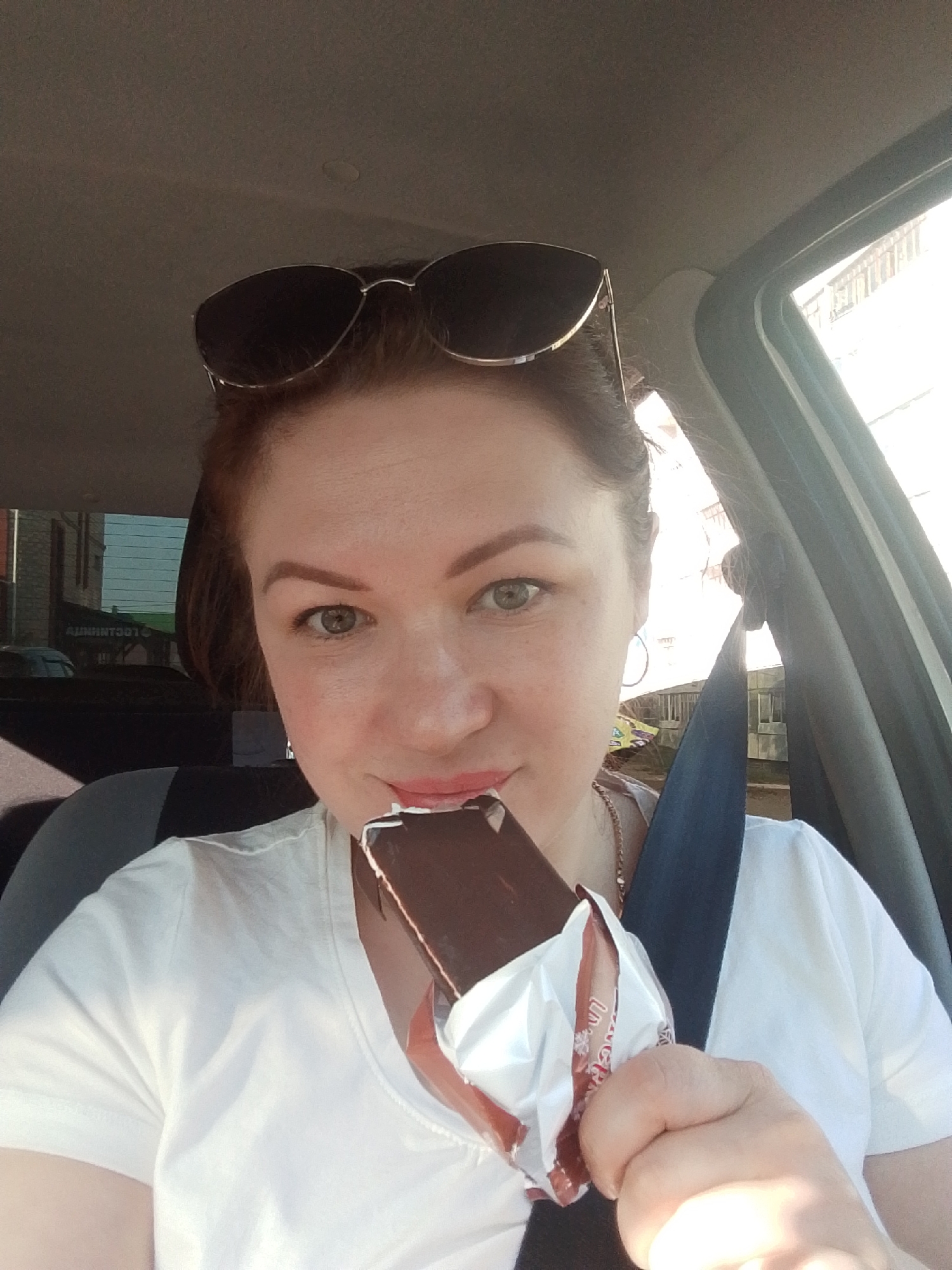A woman is shown in the driver's seat of a car. She is holding a chocolate-covered ice cream bar, partially unwrapped. She has sunglasses on top of her head and is wearing a seatbelt and a white t-shirt. The scene is brightly lit, and through the car windows, a building is visible, although the location and city of Chaikovsky, Russia, are not discernible from the image alone.