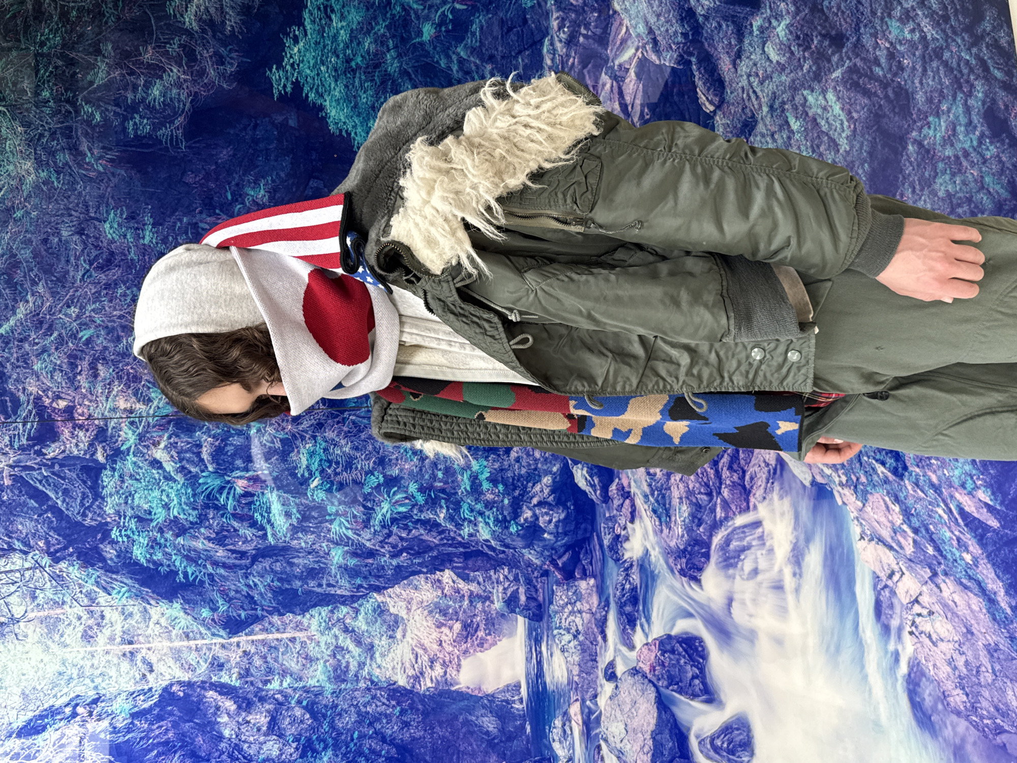 A person is standing indoors against a backdrop of a blue and white mountain landscape with a waterfall. They are wearing a grey cap, multiple scarves featuring the Japanese and American flags, a grey inner jacket with fur lining around the neck, and an outer green military-style jacket. The person's hands are visible by their side. The location is Unknown, Unknown.