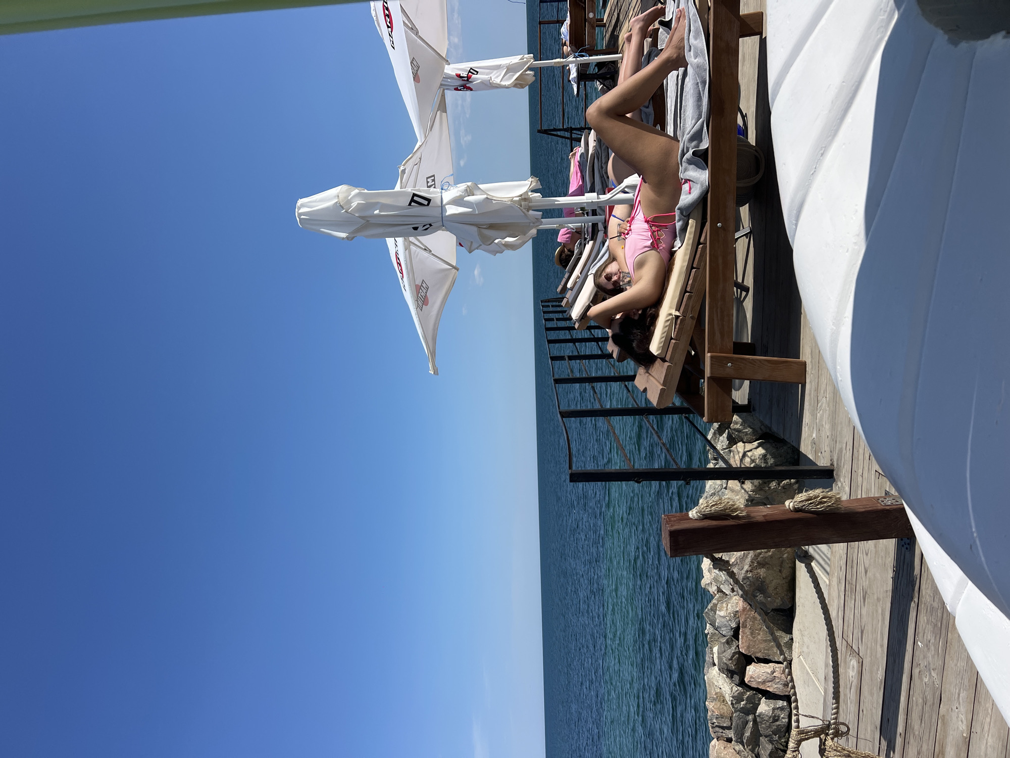 The image depicts a sunny day at a beachside location, likely in Fontanka, Ukraine, with a clear blue sky and calm ocean. In the foreground, a white boat hull is visible. Beyond this, a wooden deck area is set up with lounge chairs and beach umbrellas. Several individuals are relaxing on the lounge chairs, sunbathing. One person in the foreground is lying on a lounge chair, wearing a pink swimsuit and appearing to be holding their head. Another person is visible further back on a lounge chair, also sunbathing. Beach umbrellas, some covered, are positioned to provide shade. The ocean is a deep blue, and a rocky shoreline is visible to the left of the deck. A black metal railing separates the deck from the rocky area. The overall scene conveys a relaxed atmosphere at a seaside resort.