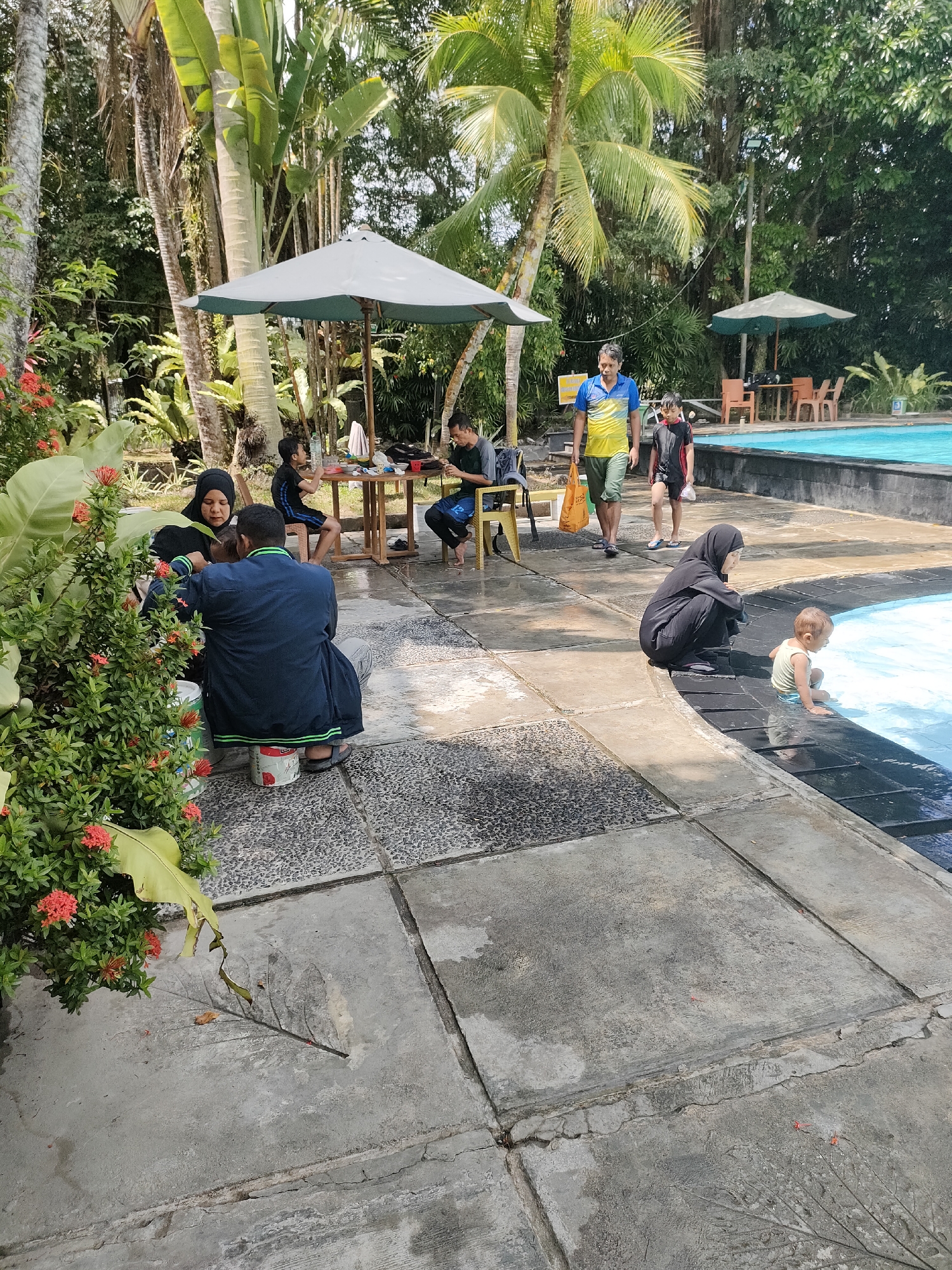 This is a medium-shot, eye-level, vertical photograph of a group of people gathered near a swimming pool. In the foreground, on the left, a woman wearing a black hijab and a green shirt is interacting with a young child, both seated on the ground. Next to them, a man in a dark blue jacket with green trim is also seated, facing away from the camera. To their right, closer to the pool, a woman in a black hijab is kneeling and looking at a baby who is sitting at the edge of the pool.
In the mid-ground, several people are seated at a wooden table under a large white umbrella. One man, wearing a blue and yellow t-shirt, is walking across the scene from left to right, carrying a bag. To his right, a young boy in a black and red swimsuit walks behind him. Further in the background, another umbrella shades a table and chairs.
The environment appears to be a tropical resort or park. Lush green foliage, including large palm trees and other tropical plants, surrounds the area. The ground is paved with large square concrete tiles. The swimming pool occupies the right side of the image, with bright blue water. The lighting suggests it is daytime, with sunlight casting shadows.