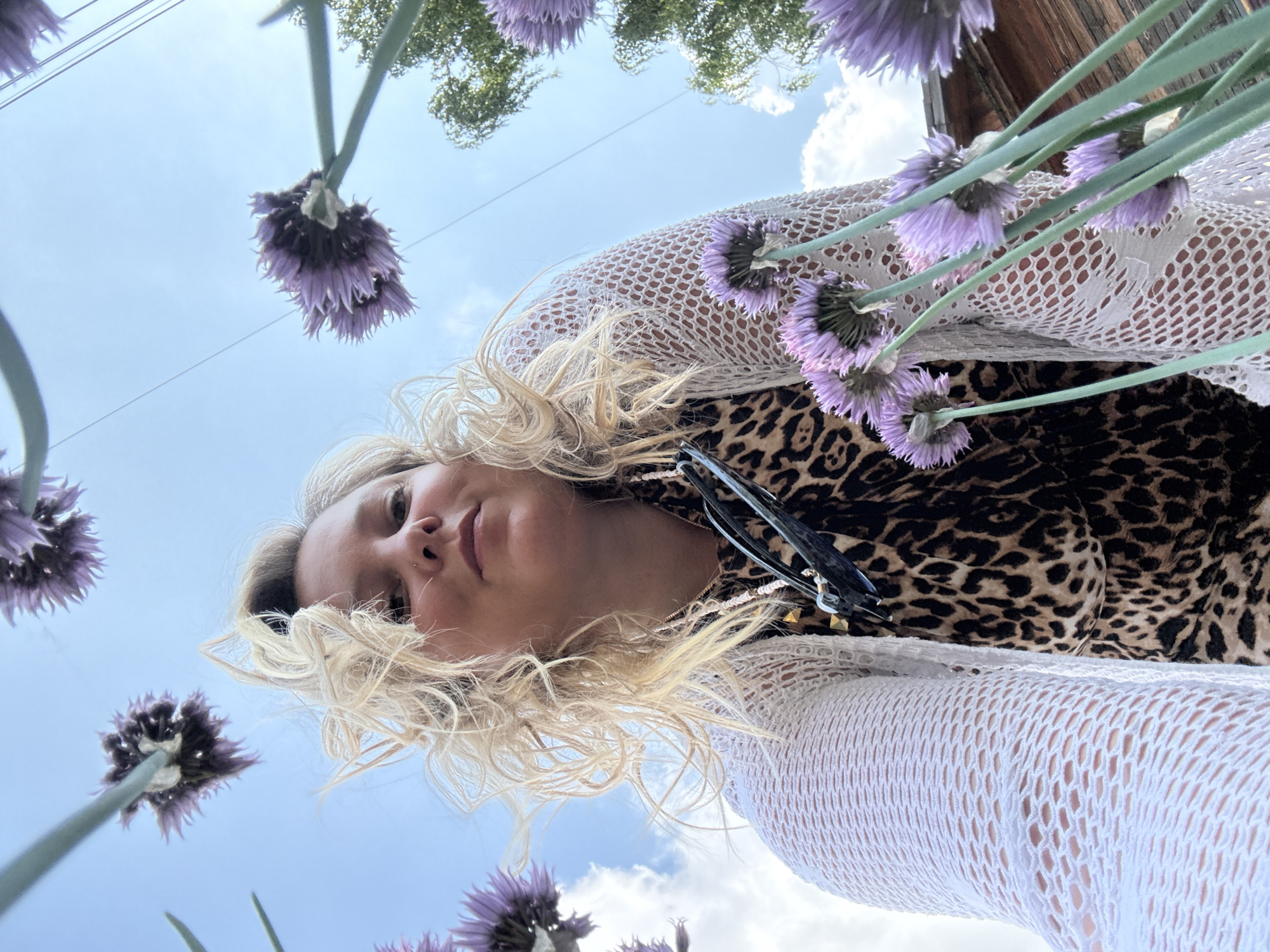 The image captures a low-angle perspective of a woman outdoors, surrounded by purple chive flowers. The woman has blonde, curly hair and is wearing a leopard-print top and a white crocheted cardigan. She is looking upwards towards the camera. The background consists of a blue sky with scattered clouds and some green trees and a wooden structure. The scene appears to be in a natural or garden setting, possibly in the city of Chaikovsky, Russia, though this cannot be definitively confirmed from the image alone.
