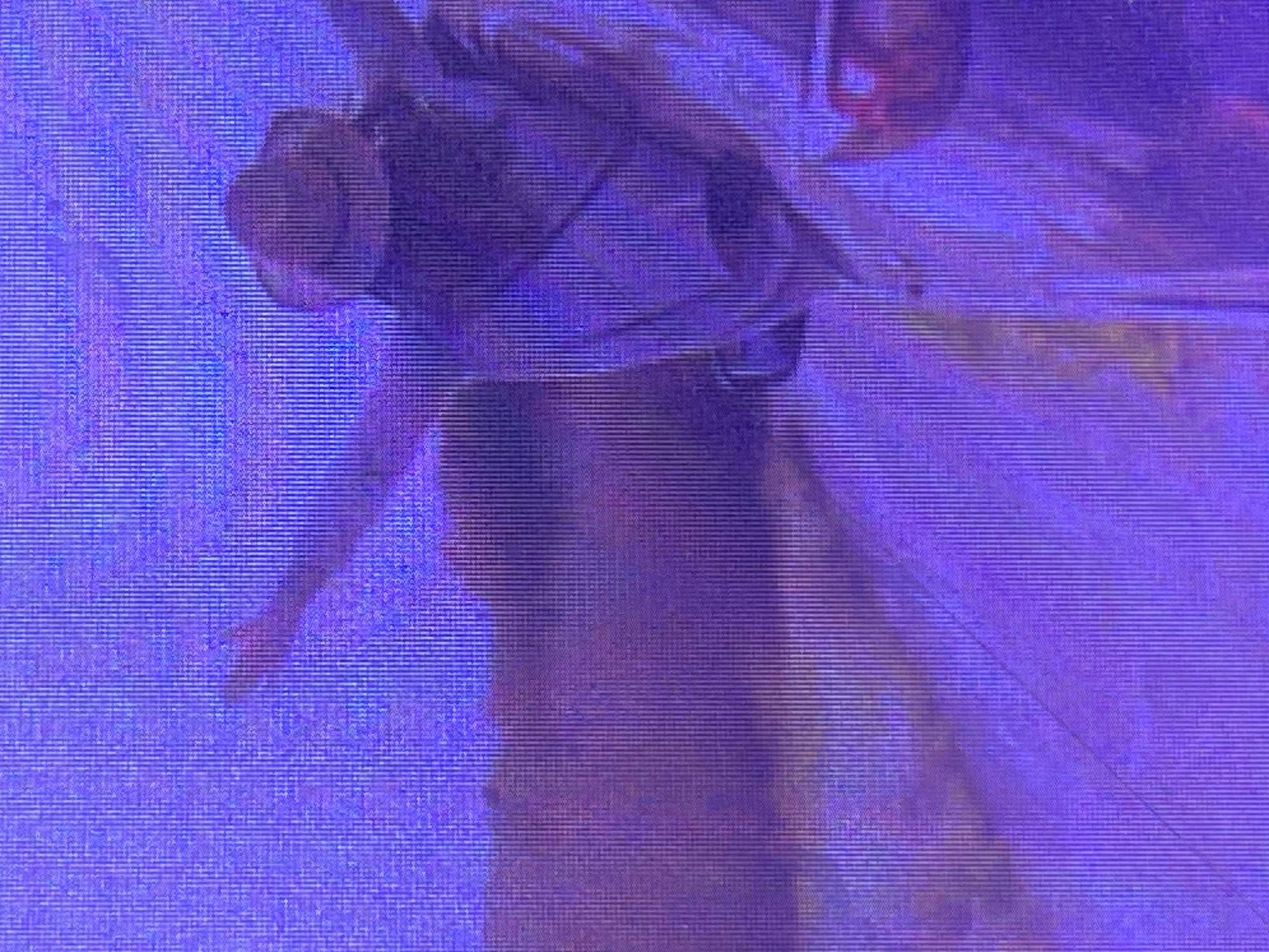 This is a digital image with a purple and blue hue, featuring a person from the waist up. The subject is facing left, with their head tilted down and their left arm extended downwards and slightly forward. The person appears to be wearing clothing that covers their torso and extends out of frame. The image has a grainy texture with visible horizontal lines, suggesting it might be a digital capture with interference or a low-resolution display. The lighting is diffused, casting a soft glow on the subject. There are no clear environmental cues to indicate the time of day, weather, or specific location within Ikorodu, Nigeria. The overall mood is somewhat obscured by the image quality, but the pose suggests a dynamic action or a moment of reflection. No text is visible in the image.