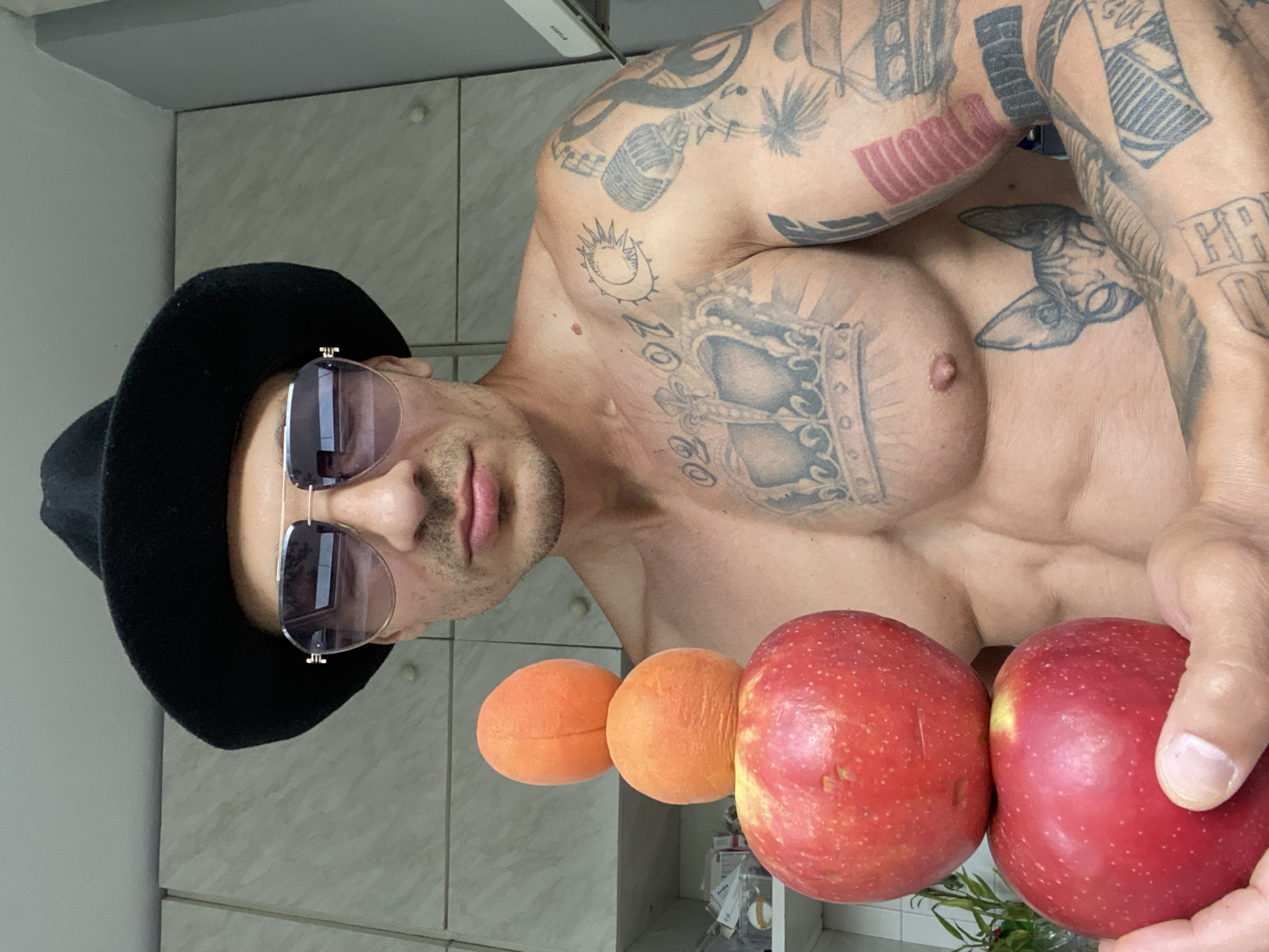 A shirtless man wearing a black fedora and aviator sunglasses stands in front of kitchen cabinets. He has a muscular physique with extensive tattoos on his chest and arms, including a crown, a crescent moon with a sun, a microphone, a cat, and various lettering. In his hands, he is holding a stack of three fruits: two red apples at the bottom and two orange apricots stacked on top. The background appears to be a domestic kitchen setting. The location is not specified in the image or provided context.