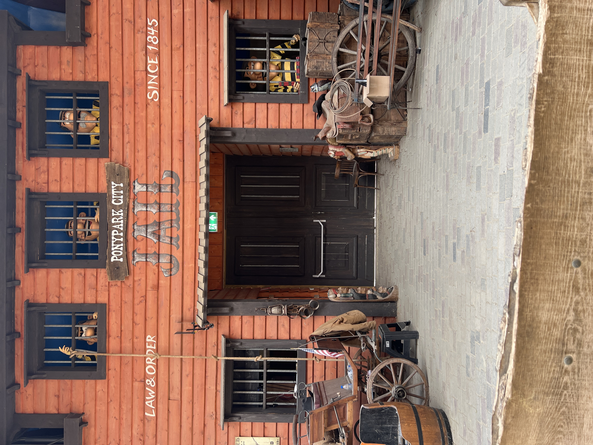 The image depicts the exterior of a wooden building with an orange facade, designed to resemble a jail. The building is located in the Ponypark City attraction in Collendoorn, Netherlands. Above the entrance doors, the words "PONYPARK CITY JAIL SINCE 1845" are visible, along with the inscription "LAW & ORDER" on the left side of the building. Three windows on the upper level of the building show cartoon figures, resembling prisoners, peering out from behind bars. The entrance features double wooden doors. In front of the building, to the left, is a horse-drawn carriage with a barrel and a wagon wheel. To the right, there are stacked wooden crates, saddles, a cowboy hat, and a wagon wheel. The ground is paved with bricks. The scene is outdoors, with clear daylight.
