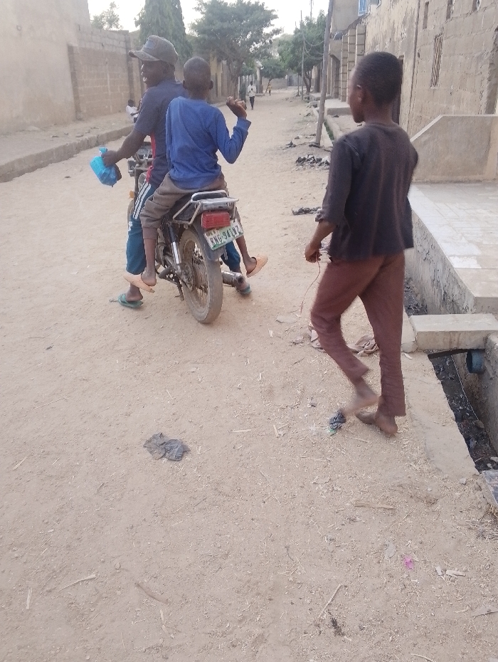 In a sandy street setting, an adult on a motorcycle carries a child as a passenger. A second child walks nearby. Buildings line the street. The motorcycle has a visible license plate. Debris litters the ground. The scene appears to be in a rural or developing area, showing everyday life.