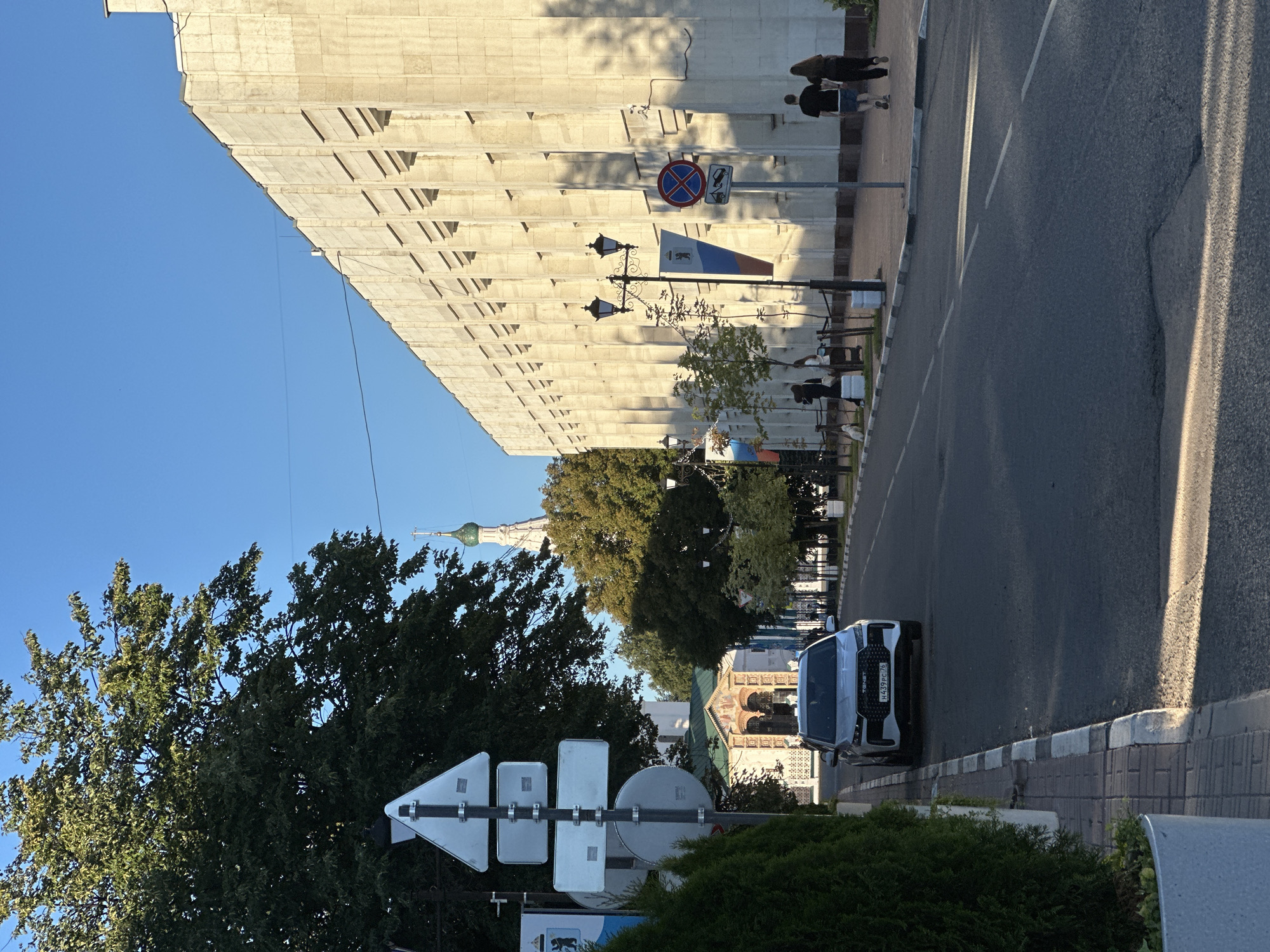 The image depicts a street scene in Yazvitsevo, Russia, bathed in the warm light of late afternoon. A modern silver car with a license plate starting with "H439 PC 76" is parked on the left side of the road, close to a sidewalk lined with manicured green bushes. The road itself is paved and has visible lane markings.
To the left, a traffic signpost with multiple triangular and rectangular signs stands prominently. Beyond the car, a building with an arched entrance and decorative facade is visible, with a Russian flag banner hanging to its right. Further in the background, the top of a white church tower with a green dome and cross pierces the clear blue sky.
On the right side of the street, a long, pale-colored building with a textured facade stretches into the distance. This building features repetitive vertical indentations and windows. A lamppost with two ornate lamps stands in front of it. Two people, a man and a woman, are walking away from the camera on the sidewalk in front of this building. Another lamppost is visible further down the street. The overall environment suggests a well-maintained urban area with architectural elements hinting at both historical and modern influences.