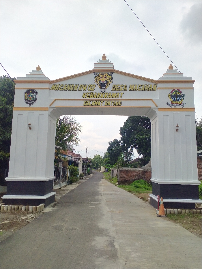 The image captures a static scene featuring a white and gold archway spanning a paved road, serving as an entrance or welcome gate to a community. No people are visible in the frame.
The archway is the dominant subject, constructed from white masonry with gold trim along its edges and decorative elements on top of its columns. At the center of the arch, a large, stylized yellow and black tiger head emblem is prominently displayed. To the left and right of the tiger head, two shields or emblems are affixed to the arch, featuring colorful designs that appear to be coats of arms or regional symbols.
Visible text on the archway reads:
* Top line: "MACANAN RW 02" (likely a neighborhood or sub-district name and number).
* Middle line: "DESA MACANAN" (Village of Macanan).
* Next line: "KEBAKKRAMAT" (referring to Kecamatan Kebakkramat, the sub-district).
* Bottom line: "SELAMAT DATANG" (Welcome).
The road beneath the archway is paved, appearing to be asphalt or concrete, extending into the distance. It is relatively narrow, suggesting a village or residential street. Along both sides of the road, various houses and structures are visible, mostly single-story with some fencing and greenery. The architectural style appears typical of Indonesian rural or semi-urban dwellings. Lush green trees and foliage are scattered throughout the scene, particularly on the left and right sides of the archway and further down the road, indicating a verdant environment.
The sky is overcast, suggesting a cloudy day or a time of day when the sun is not directly overhead, such as morning or late afternoon. The overall lighting is soft and diffused. An orange traffic cone is placed near the base of the right archway pillar. The setting is clearly a residential area within Kecamatan Kebakkramat, Indonesia, marked by the welcoming gate.