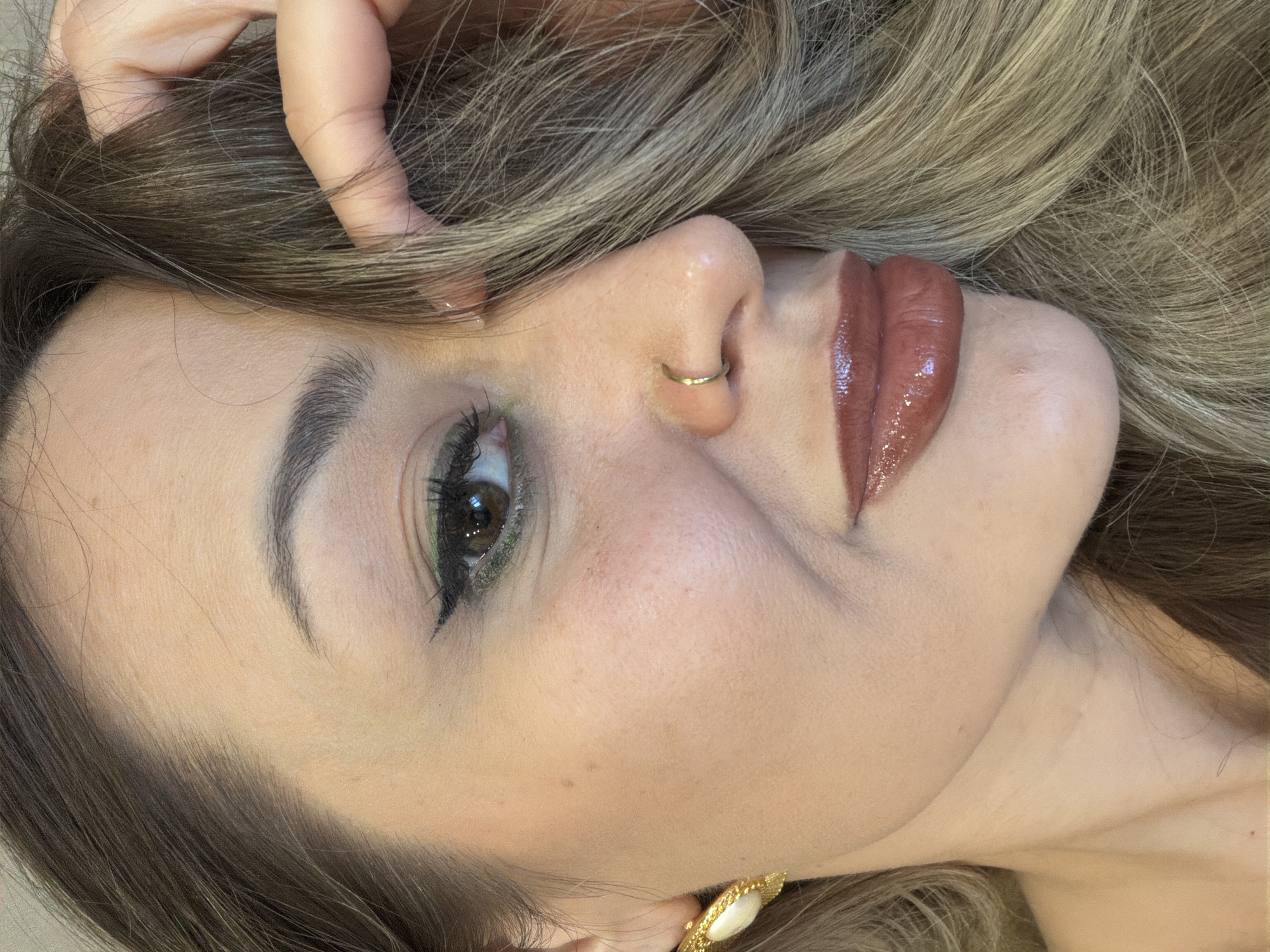 The image is a close-up, angled portrait of a woman. She has blonde and brown hair, fair skin, and is wearing makeup including eyeliner and lipstick. A gold ring is visible in her left nostril. Her left hand is positioned in her hair. An earring is visible. The location is not identifiable as Krasnoyarsk, Russia.