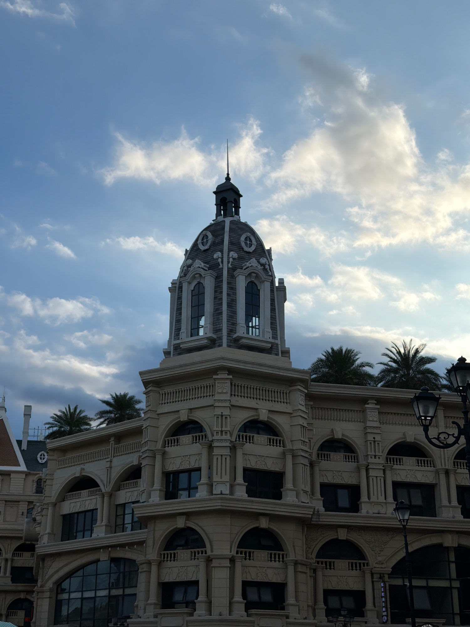 The image shows a building with a dome-shaped tower, possibly a part of the Vinpearl Land complex in Nha Trang, Vietnam. The architecture appears ornate, with classical elements like columns and arched windows. The building is made of light-colored stone or concrete. Palm trees are visible behind the building, suggesting a tropical or subtropical location. The sky above is blue with scattered clouds, indicating daytime. There are lampposts in the foreground, some of which appear to be ornate streetlights. No people are visible in the image. The overall scene suggests a resort or entertainment complex.