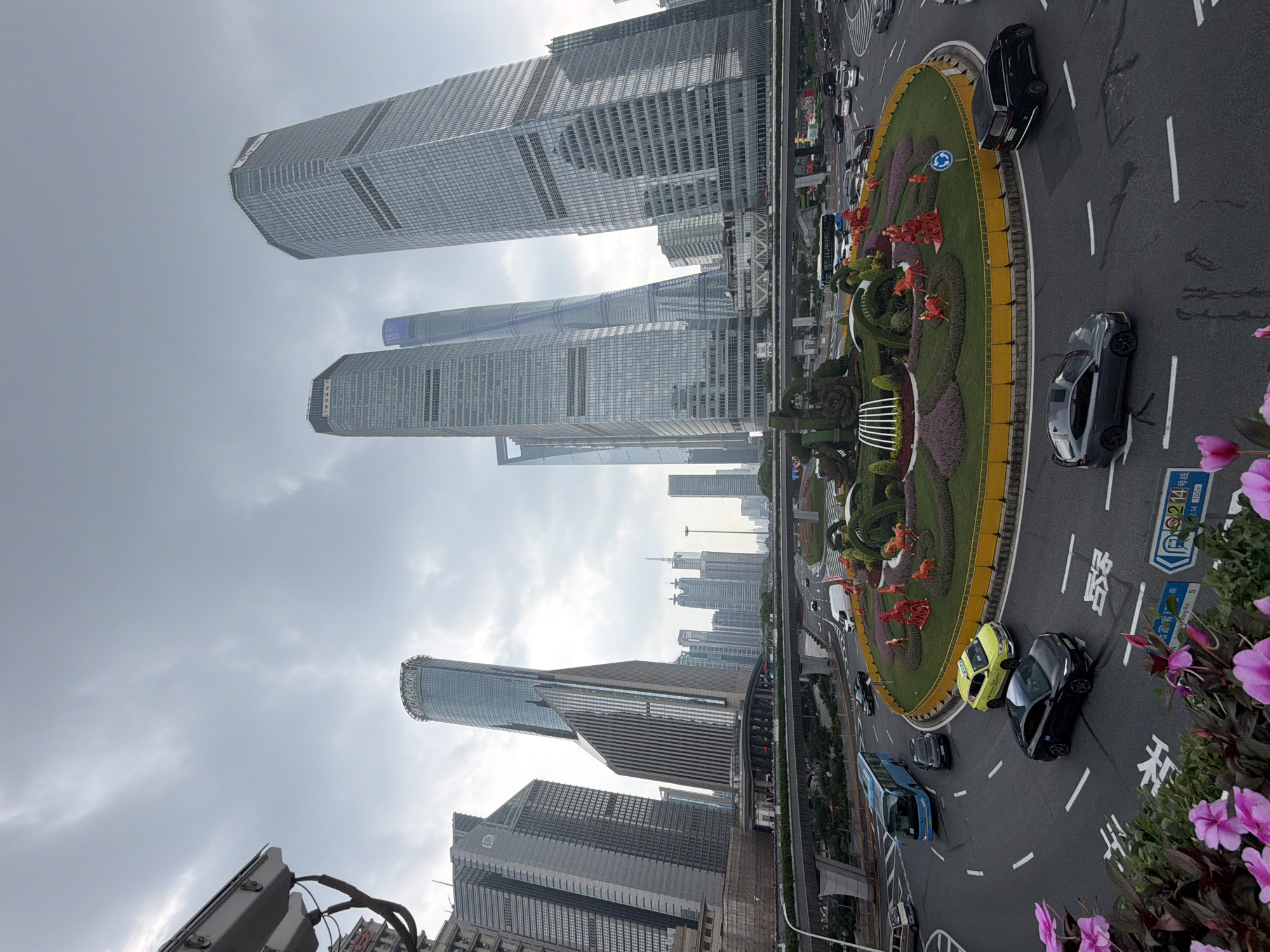 The image depicts a wide-angle view of a busy intersection in Pudong, China, dominated by modern skyscrapers. In the foreground, a circular roundabout features elaborate floral arrangements and decorative red horse sculptures. Several vehicles, including a blue bus, a yellow taxi, and black cars, are navigating the roundabout. Overhead, an elevated roadway with traffic can be seen. The background is filled with towering glass-fronted skyscrapers, indicative of a major urban center. The sky is overcast, suggesting cloudy weather. The overall scene captures the dynamic urban landscape of Pudong, characterized by its impressive architecture and active street life.