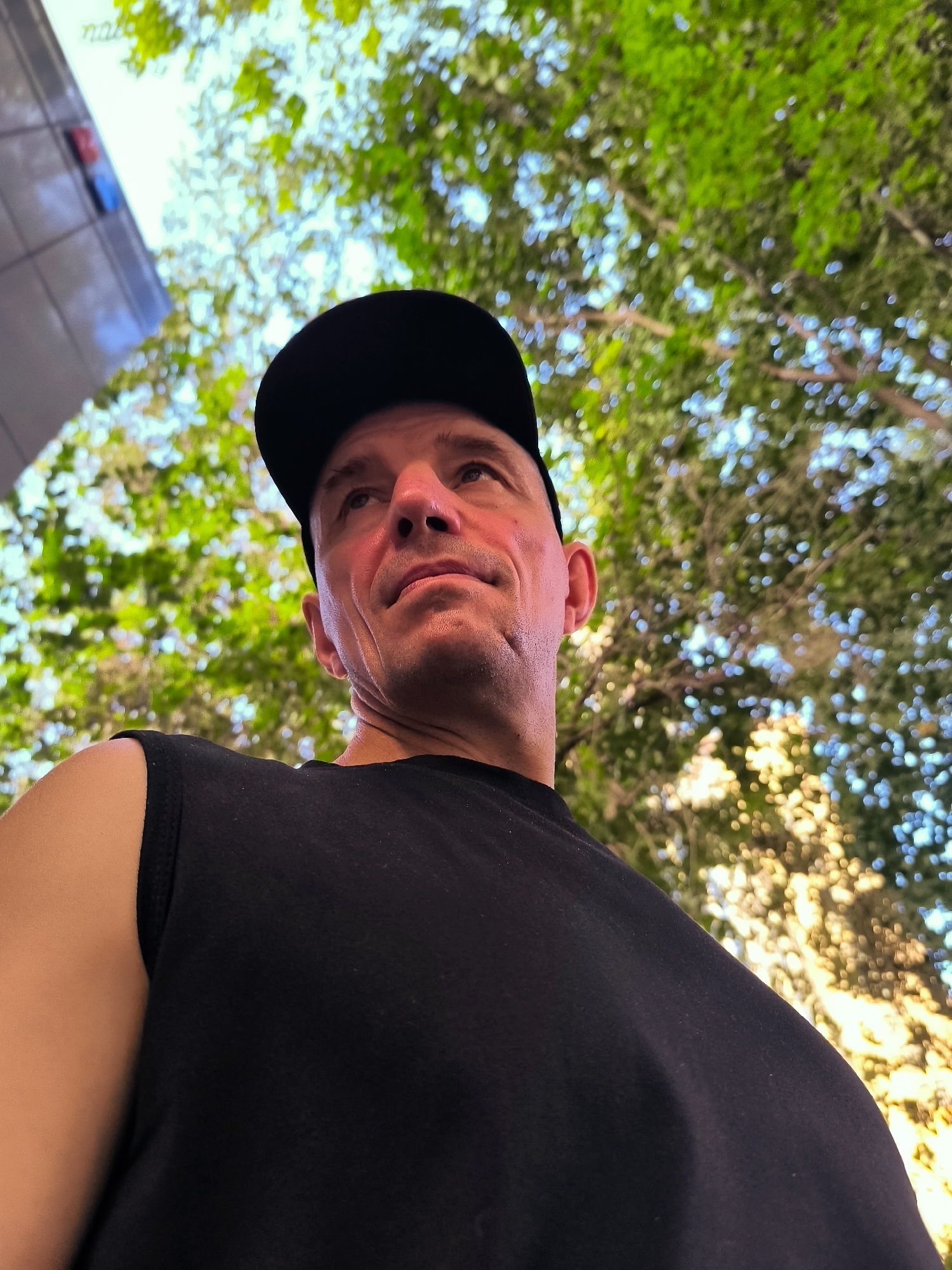The image is a low-angle shot of a man wearing a black baseball cap and a black sleeveless shirt. He is looking upwards and to his right. Behind him, out of focus, is a building with colored panels and the word "natr" visible on it, and lush green foliage. The lighting suggests it is daytime. The man's face is partially illuminated by sunlight, and his skin appears slightly reddish, possibly from the sun. The overall impression is of a casual outdoor setting, possibly in an urban park or near a modern building. There is no information in the image that can identify the location as Ürümqi, China.