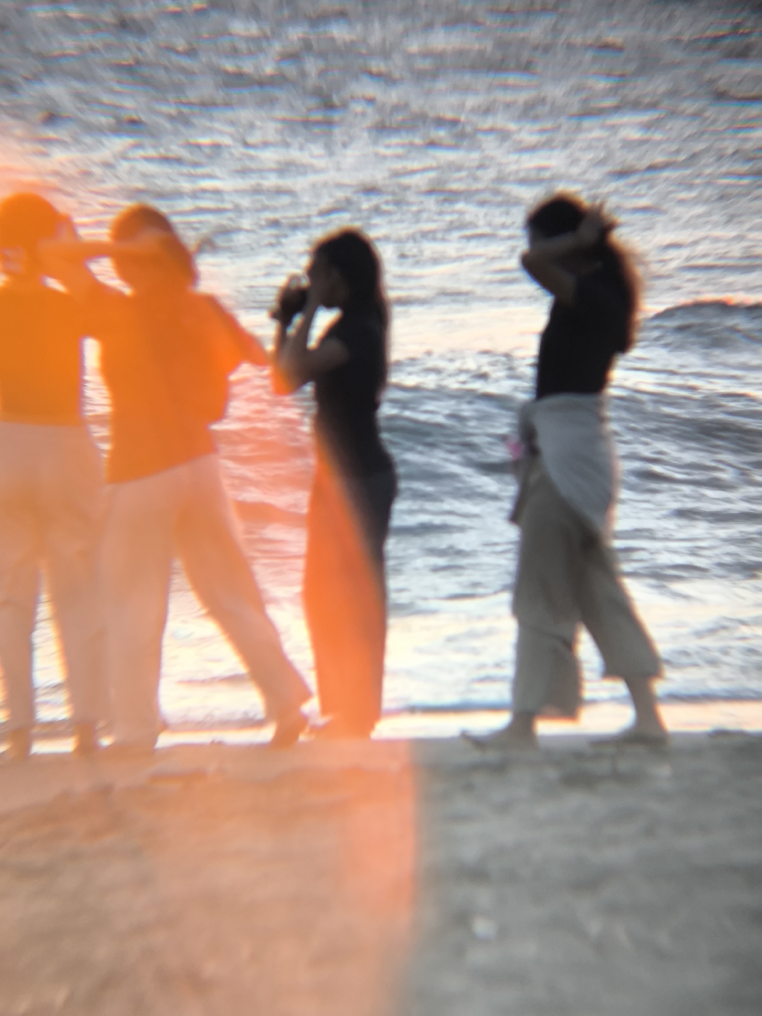 The image, blurry, depicts four individuals standing on a beach in North Kuta, Indonesia. They stand on the sand near the water's edge. The person third from the left is drinking. One person has their hands in their hair. The ocean is visible in the background. The scene seems to be taking place during sunset.